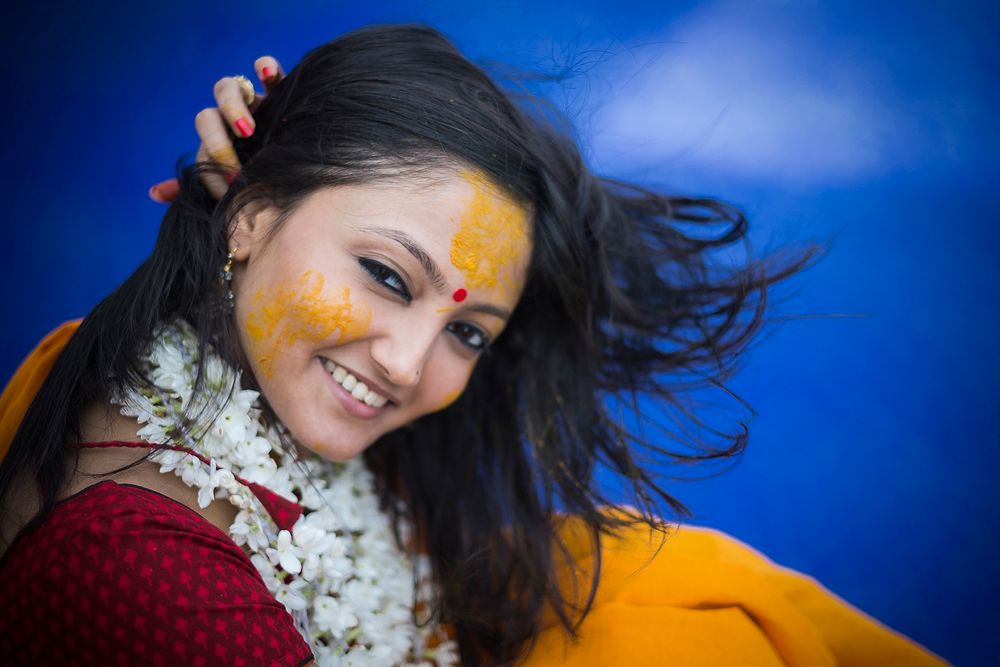 Person wearing red dress and floral garland smiles against blue background with yellow turmeric on face.