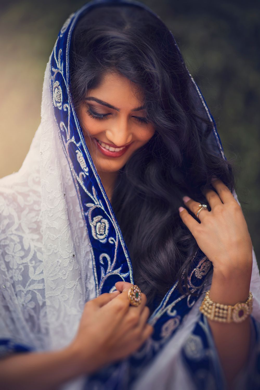 Woman wearing an embroidered blue and white dupatta with traditional jewelry smiles while adjusting her hair.
