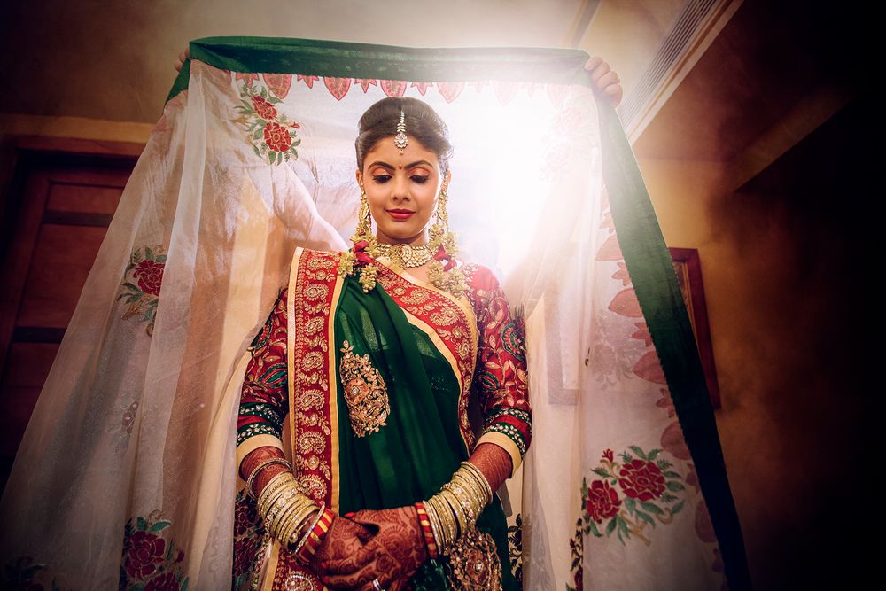 Traditional South Asian bride wearing an ornate green and red bridal lehenga with gold embroidery stands in soft window light.