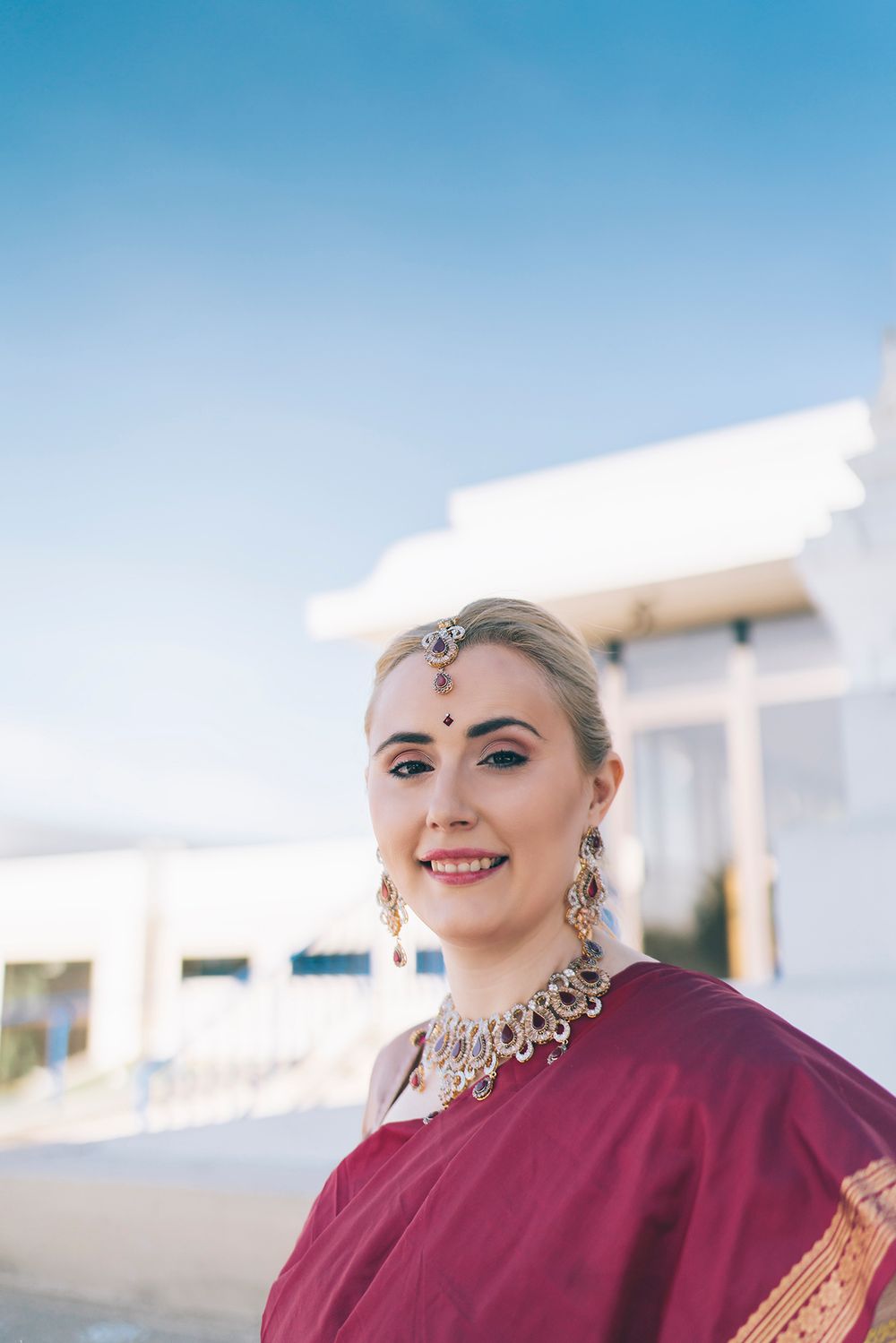 A person wears traditional Indian jewelry and a burgundy outfit while posing outdoors against a white building.