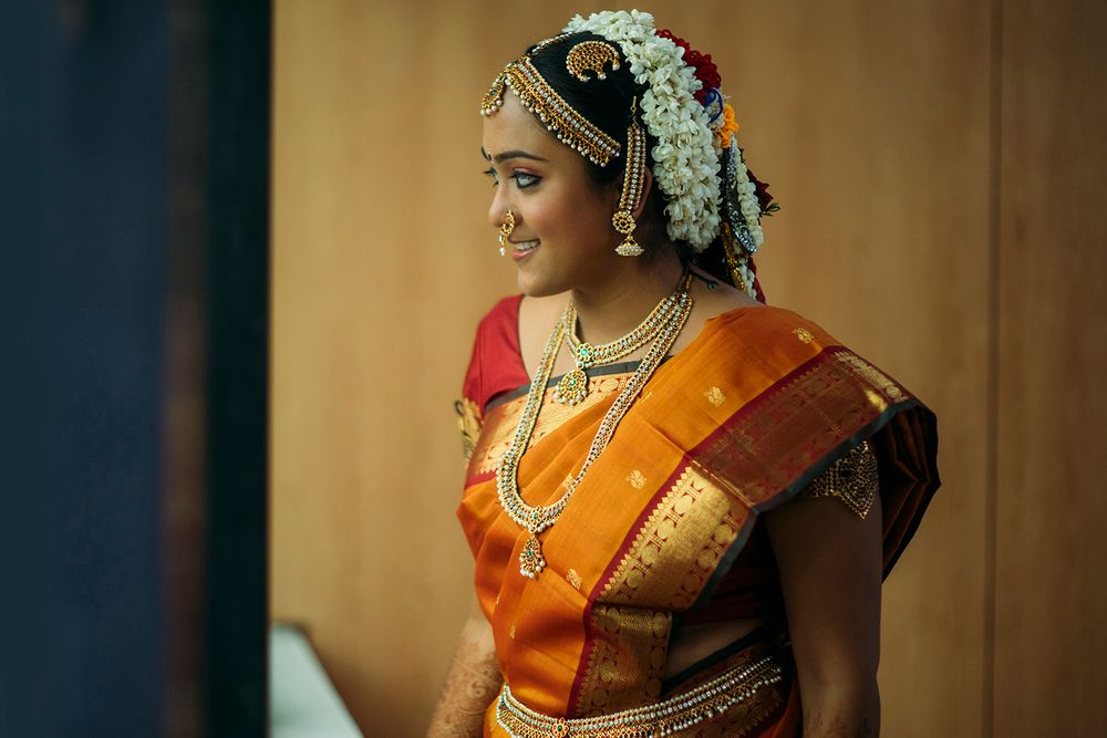 A woman wearing traditional South Indian bridal attire in orange silk saree with gold borders and floral hair accessories smiles.