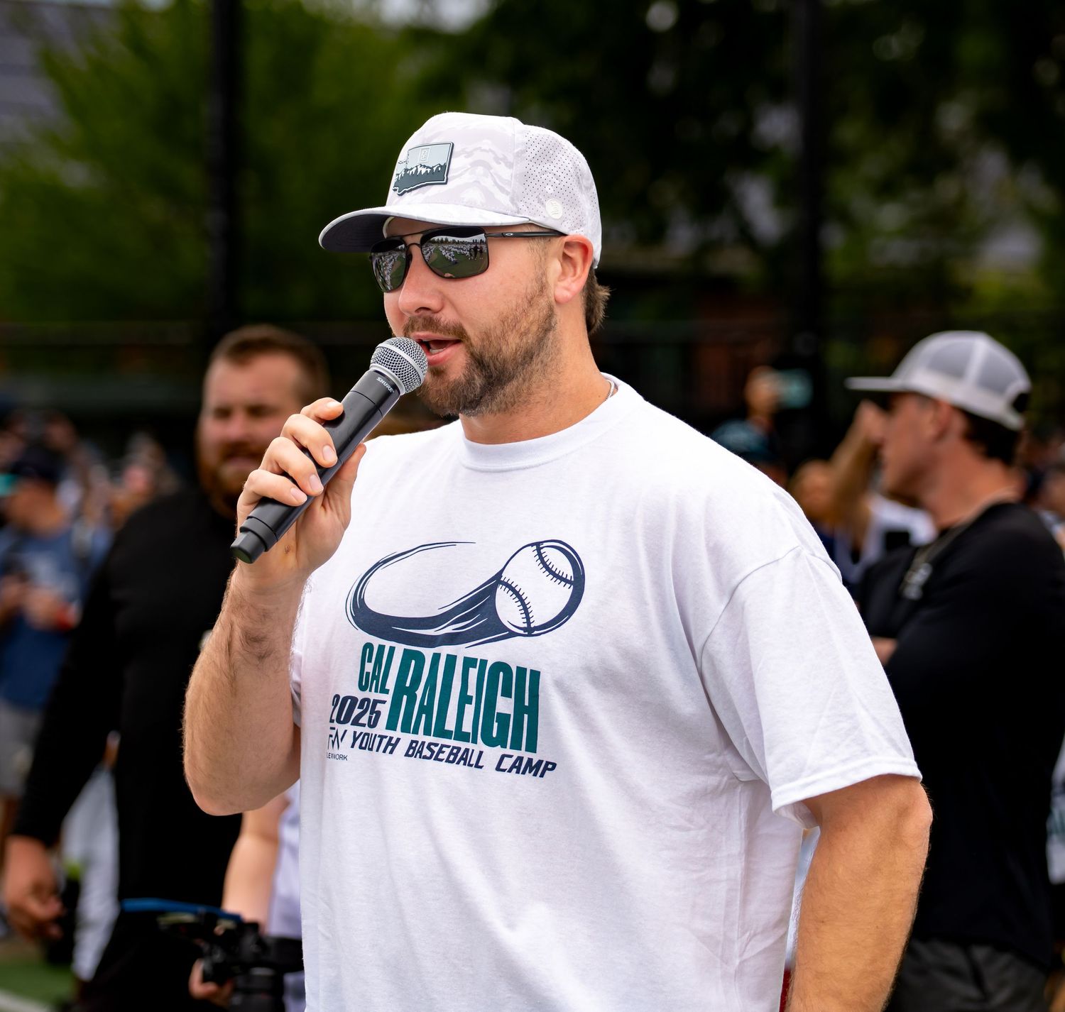 Man in Cal Raleigh Youth Baseball Camp shirt speaks into microphone at outdoor baseball event.
