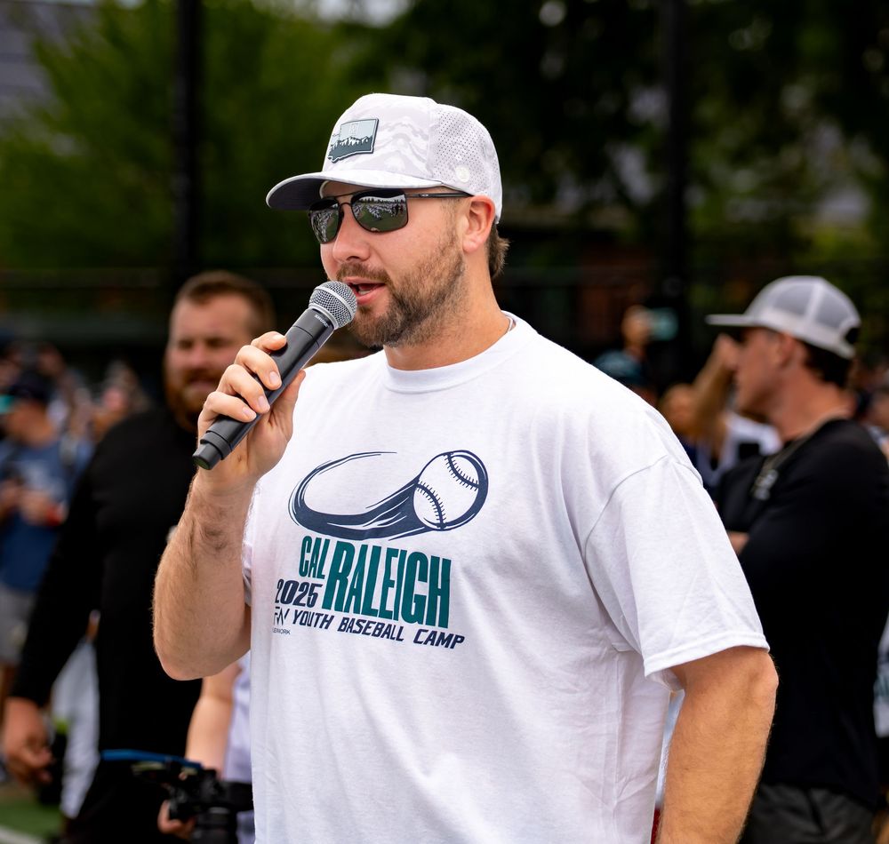 Man in Cal Raleigh Youth Baseball Camp shirt speaks into microphone at outdoor baseball event.