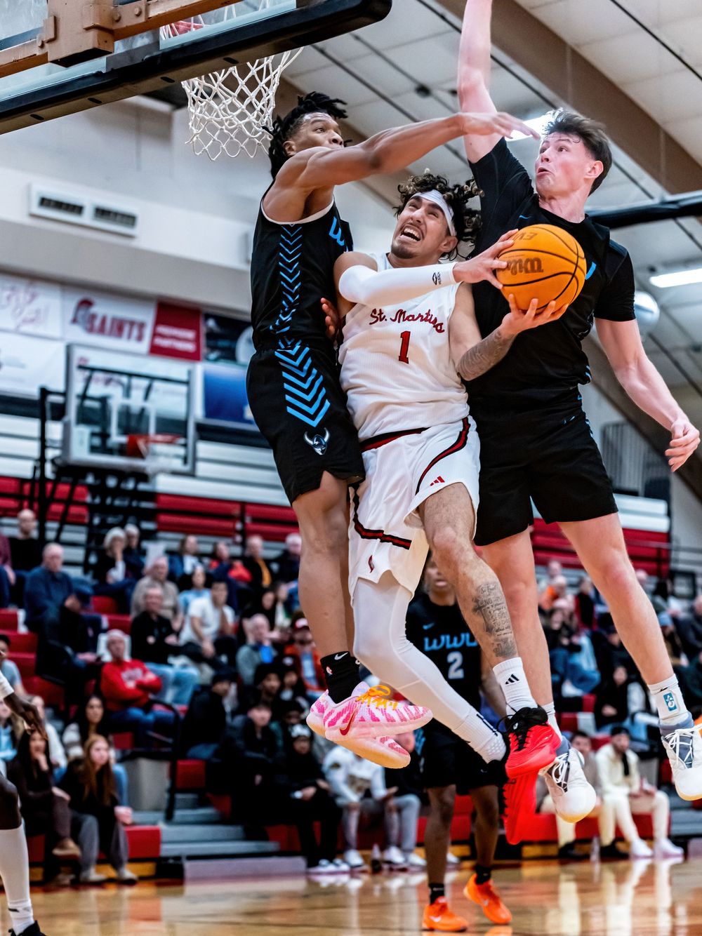 Basketball player in white jersey drives to hoop between two defenders during an indoor high school game.