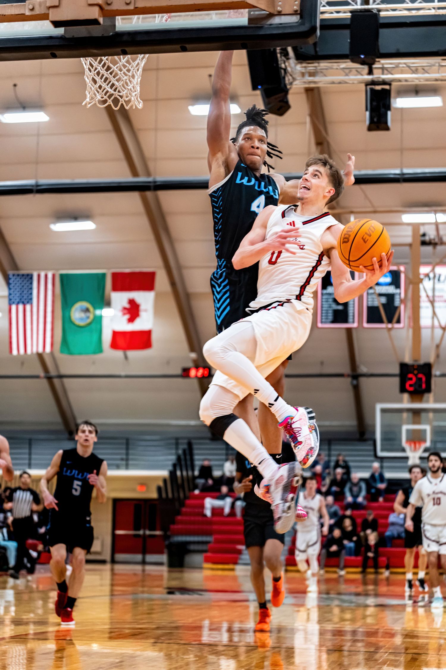A basketball player in white #0 drives past a defender for a layup in a packed indoor gymnasium.