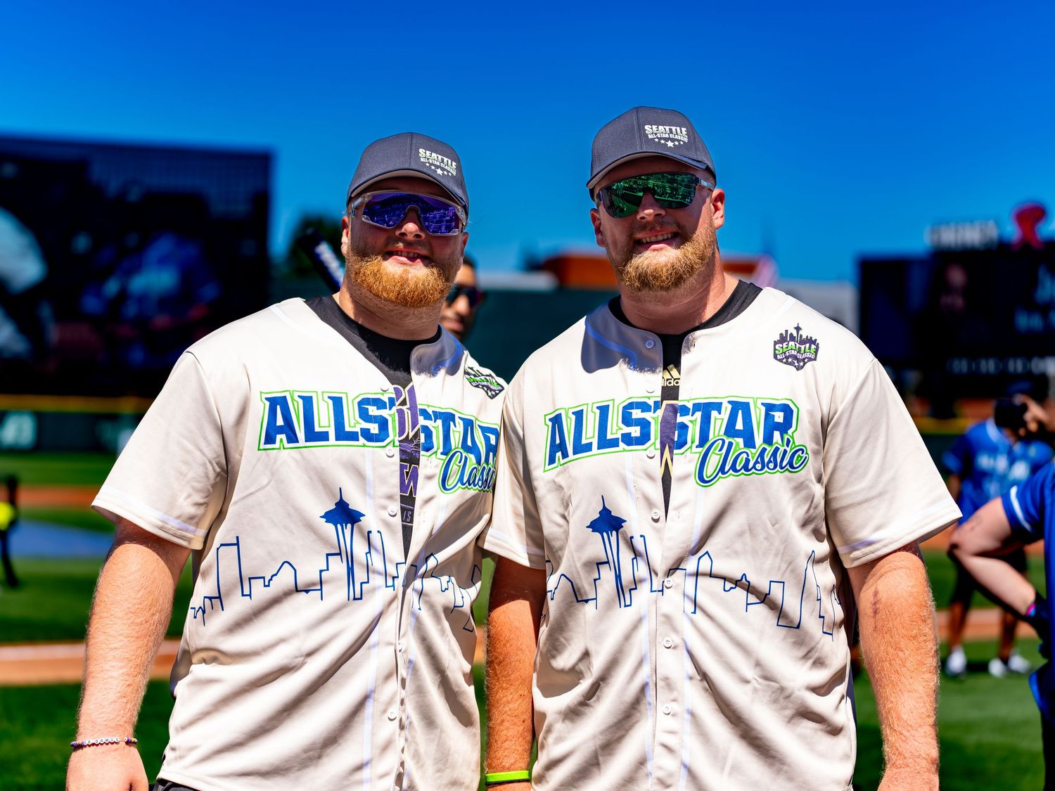 Two men wearing All Star Classic baseball jerseys and caps pose together on a sunny baseball field.