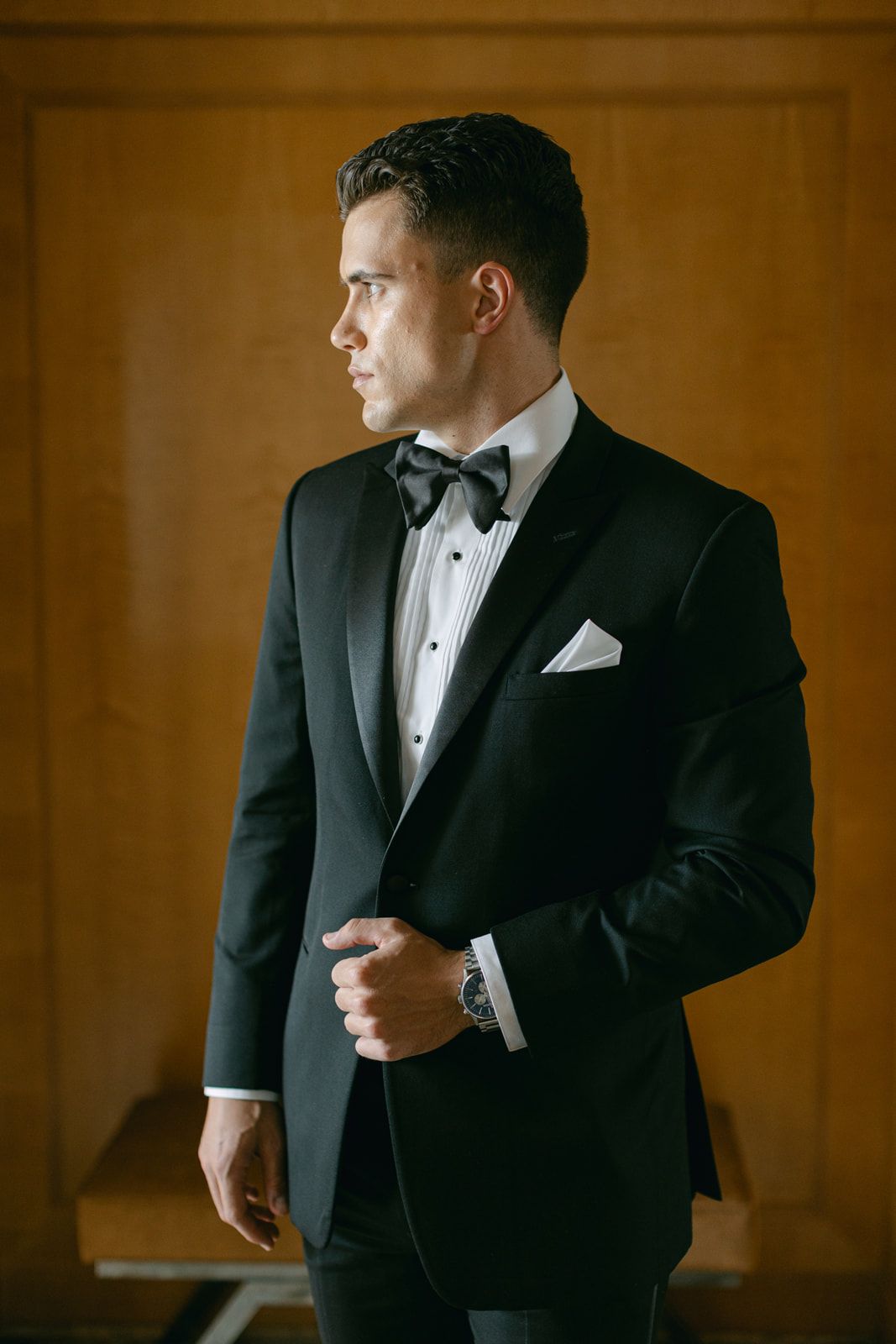Classic black tuxedo with bow tie and white pocket square photographed in elegant profile against warm brown background.