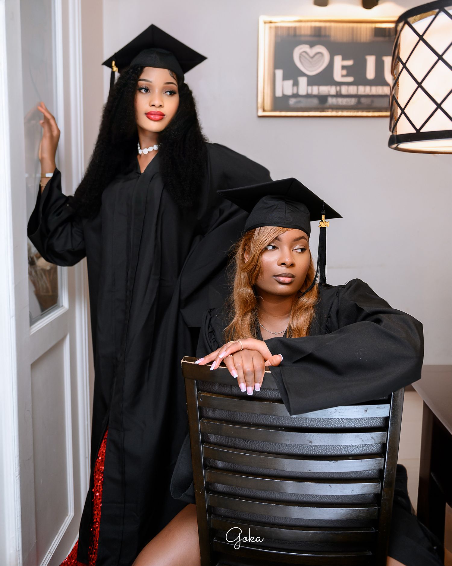A graduate poses in a commencement cap and gown on a dark chair against a studio backdrop for portraits.