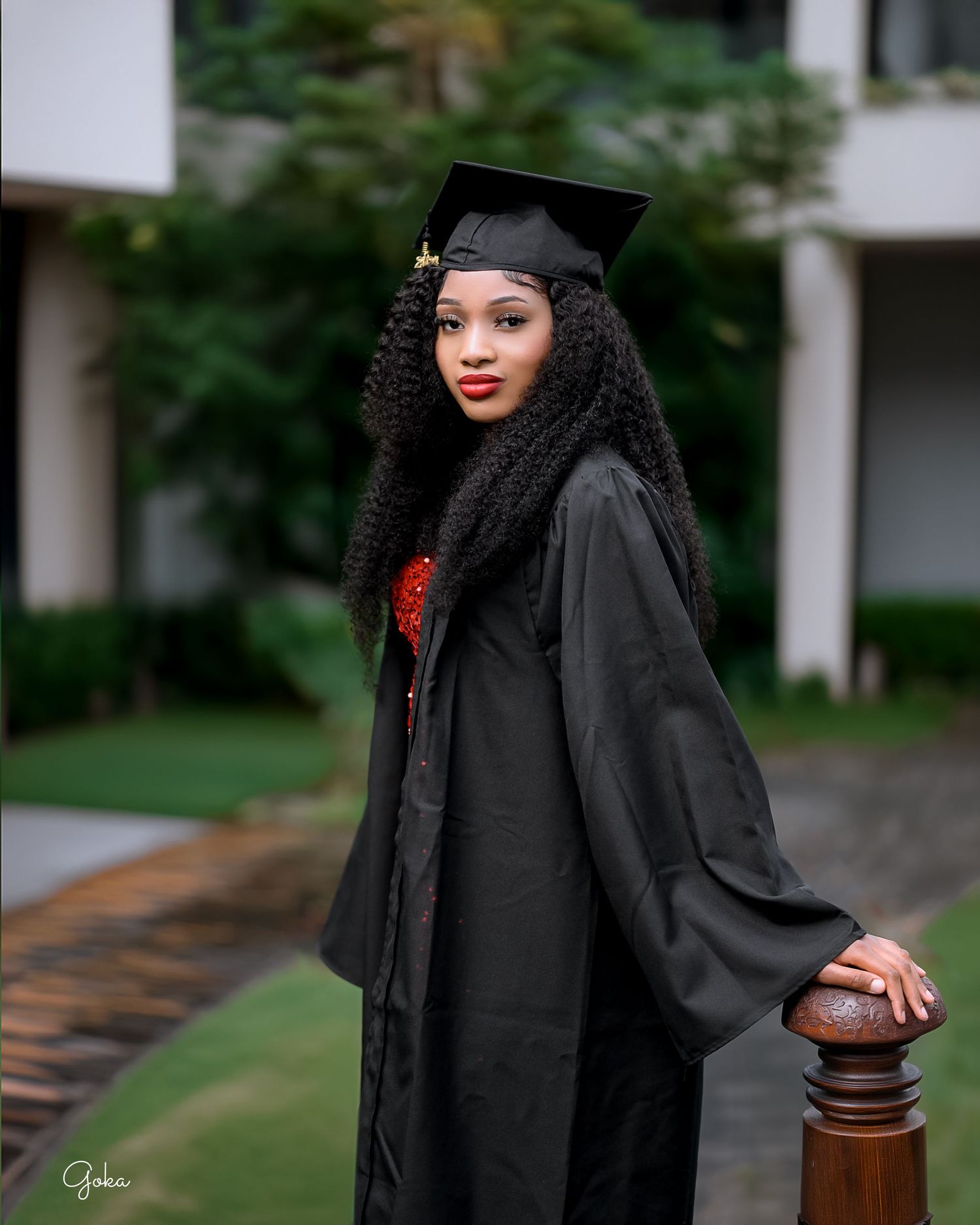 Graduate in black academic regalia and cap poses elegantly beside a wooden railing on graduation day.