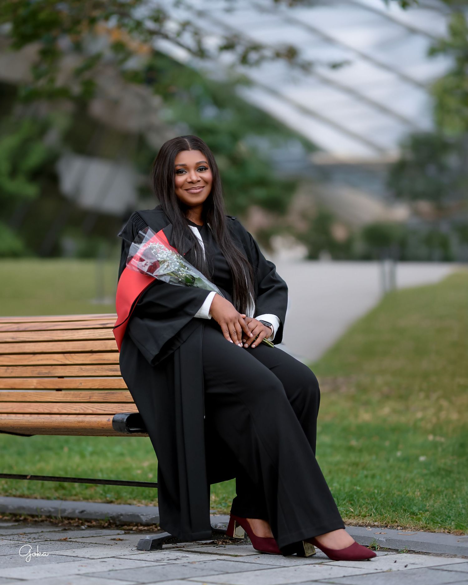 Graduating student in black cap and gown sits on wooden bench outdoors holding flower bouquet and smiling.
