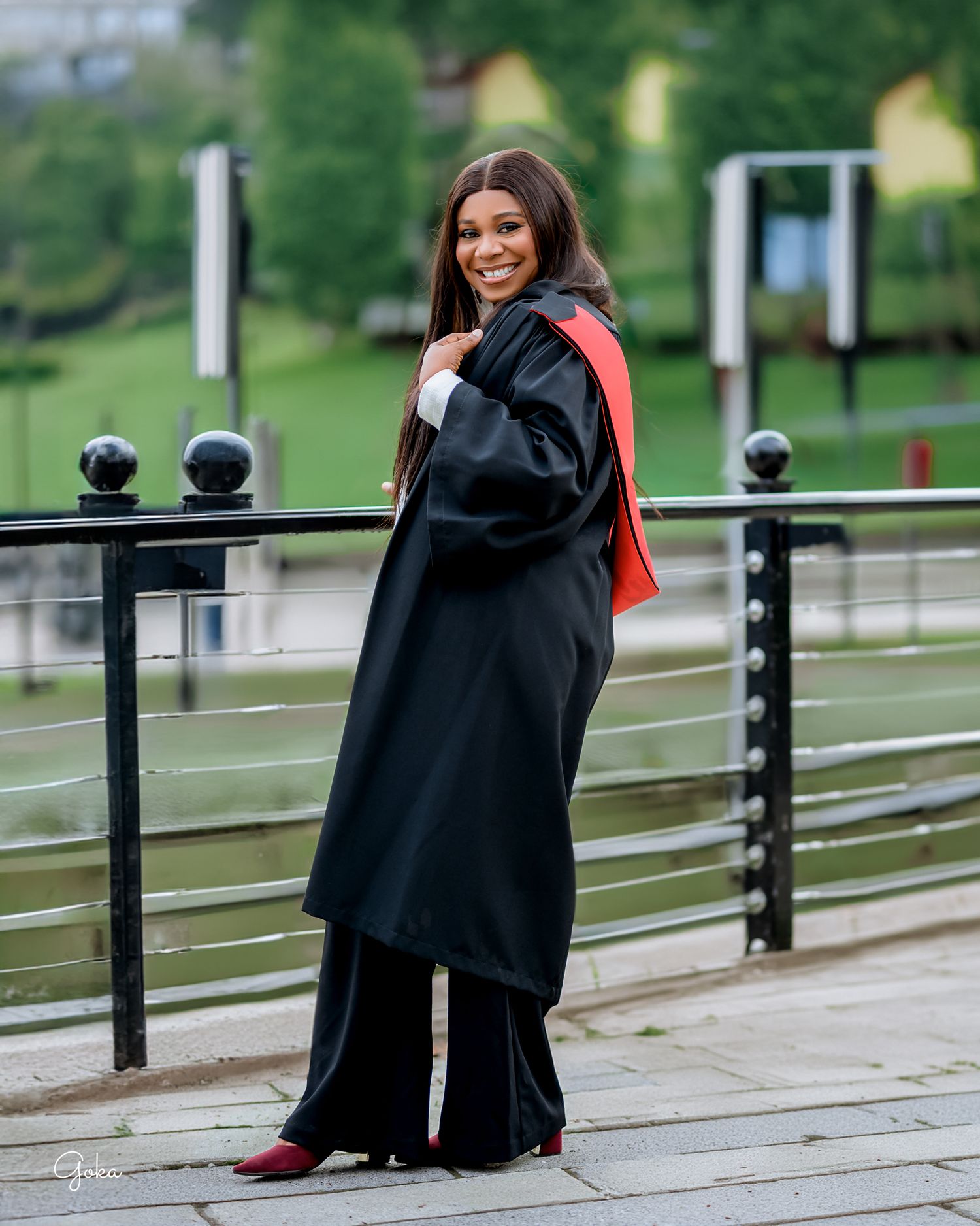 Graduate in black academic gown and red stole stands on a bridge railing during a photo session outdoors.