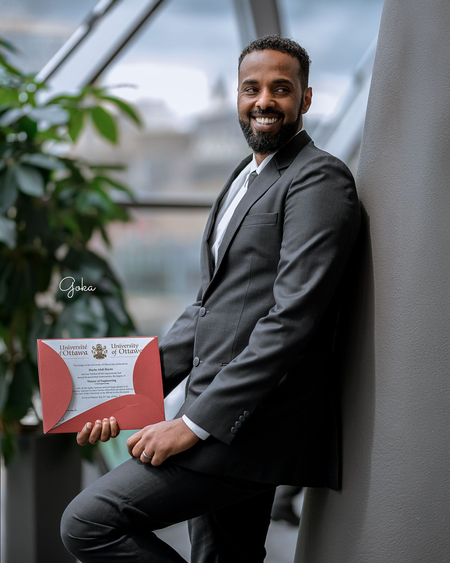 Professional in a dark suit holds a red diploma while posing confidently against an interior background with plants.