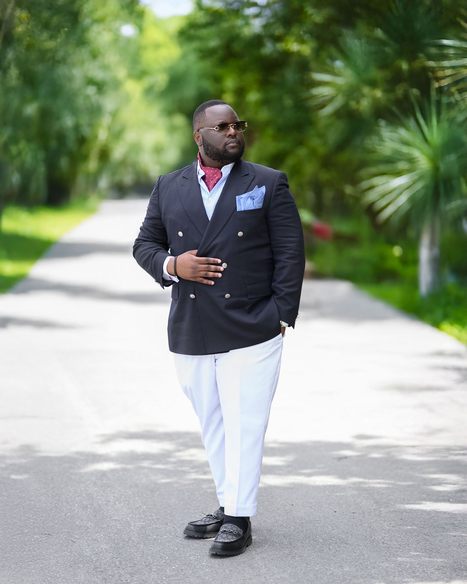 Stylishly dressed figure in a navy double-breasted blazer and white pants poses confidently on a palm-lined pathway.