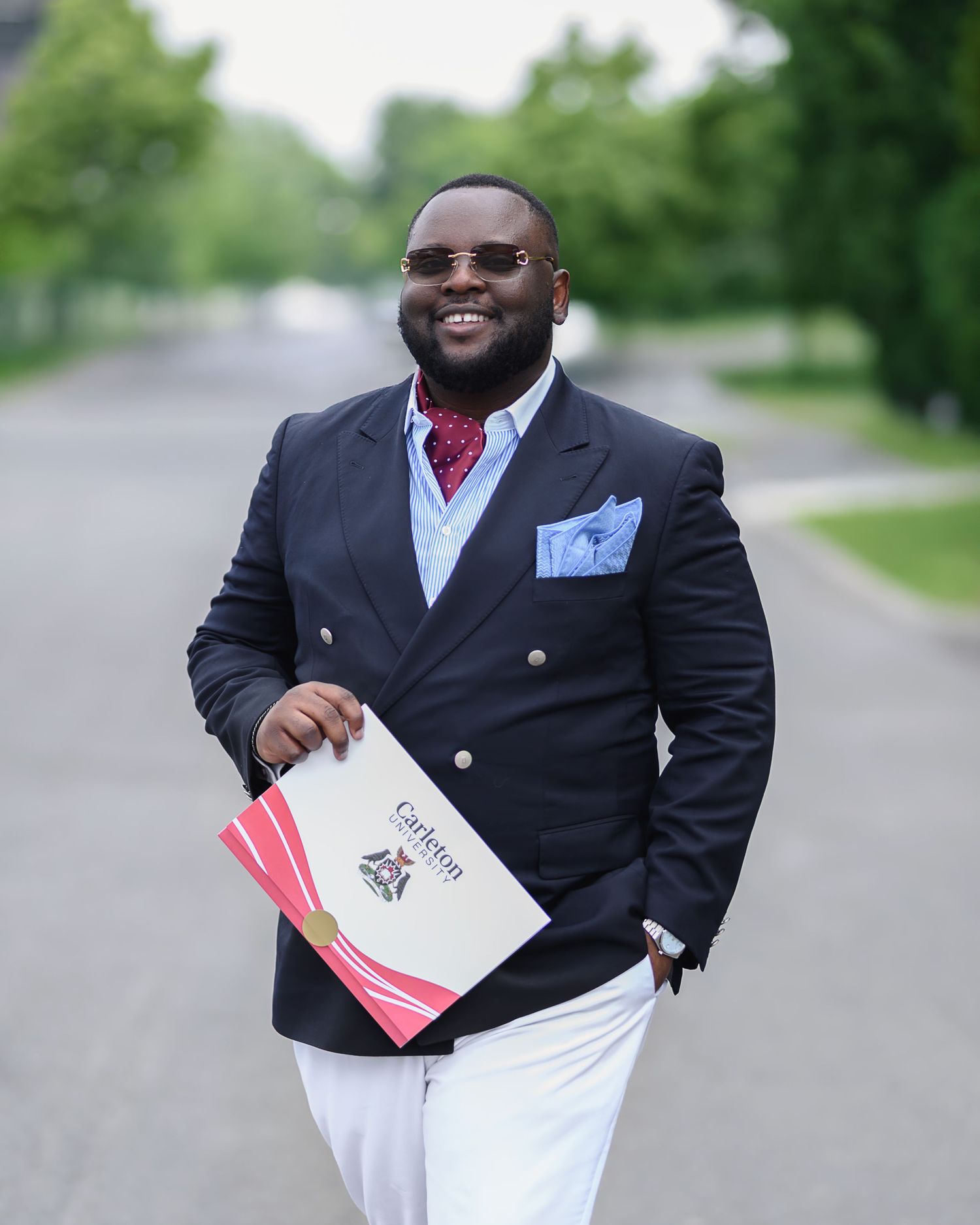 A well-dressed professional in a navy suit and white pants holds paperwork while standing on a tree-lined street.
