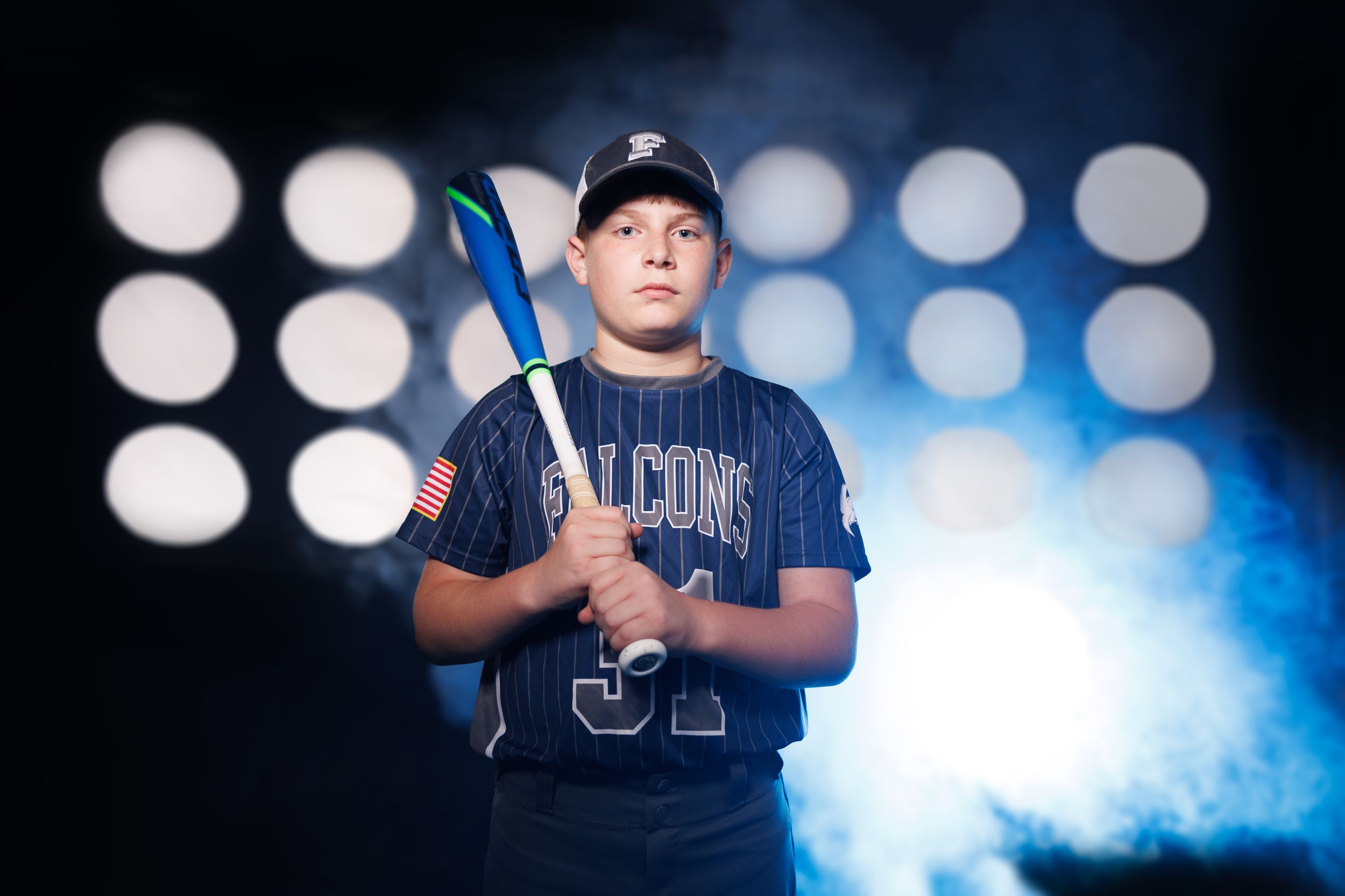 Youth baseball player poses with bat against dark background with stadium lights.