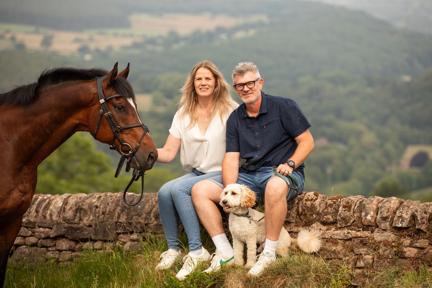Someone wearing a white patterned top lovingly strokes a brown horse in a sunny outdoor setting.