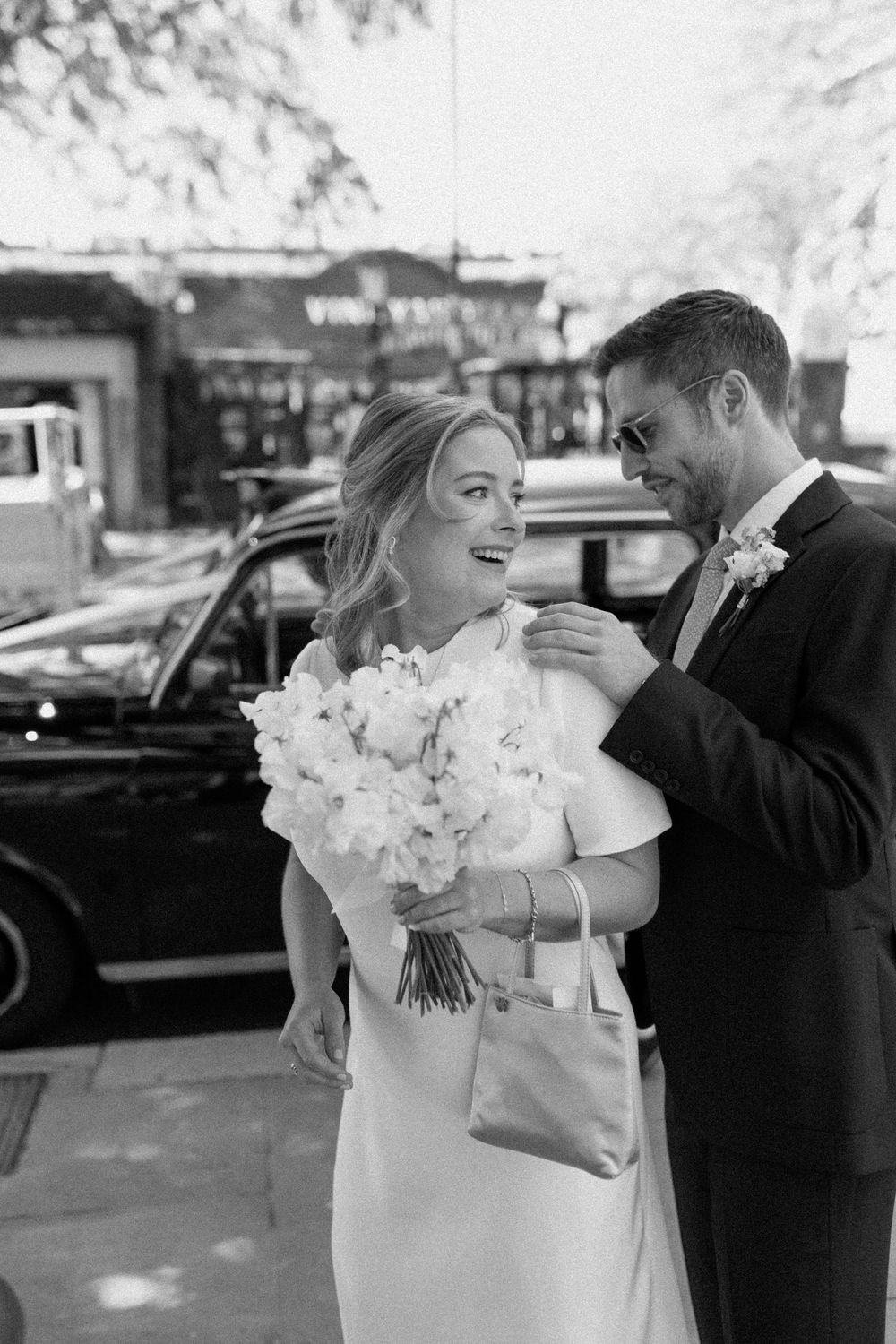 Black and white photo of a joyful moment captured on a wedding day with a gorgeous white bouquet and London cab in background.