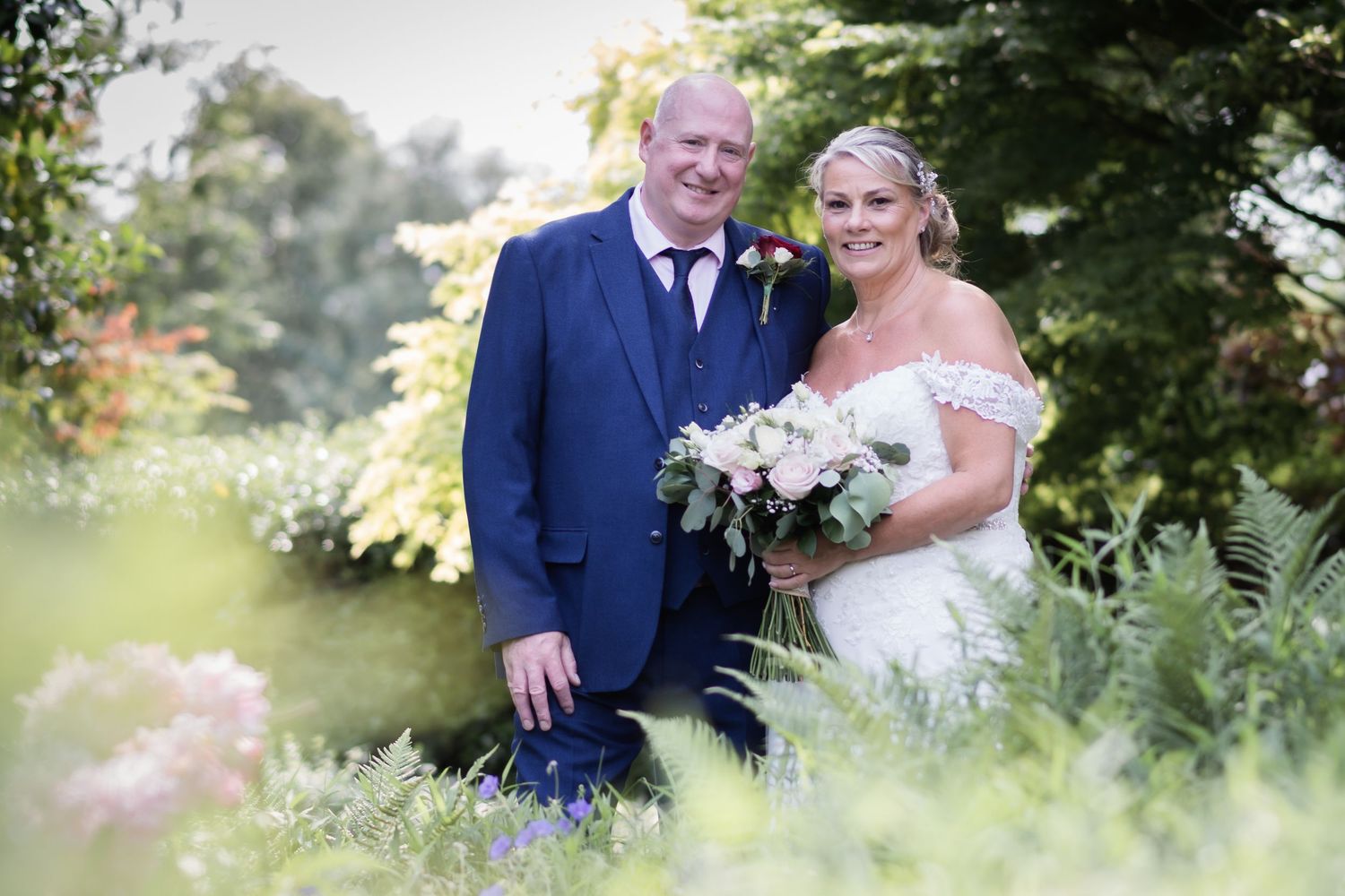 A couple poses in a lush garden setting on their wedding day, with greenery and ferns surrounding them.