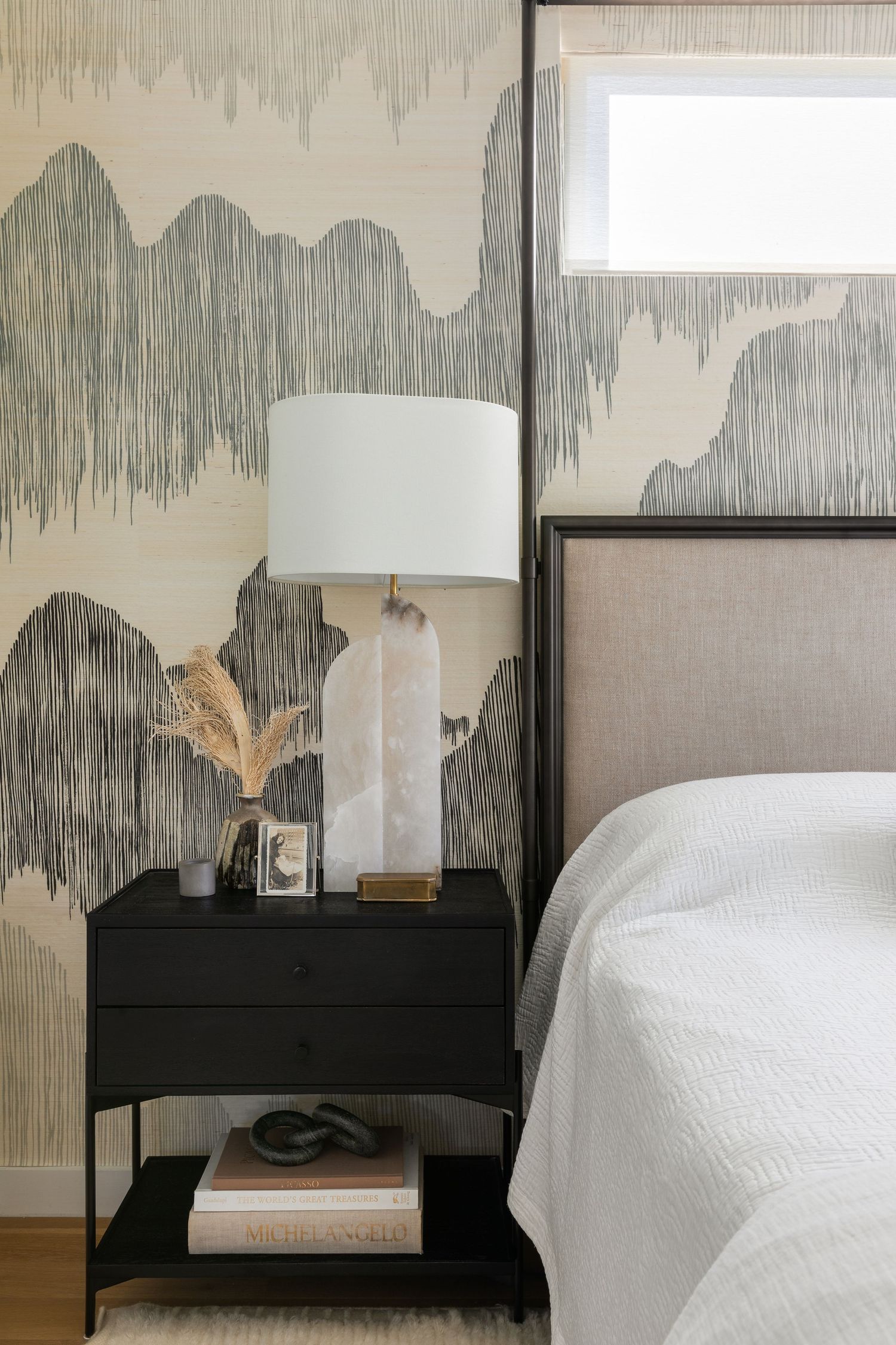 Stylish bedroom interior with abstract gray wallpaper, black nightstand, white table lamp, and partial view of white bedding.