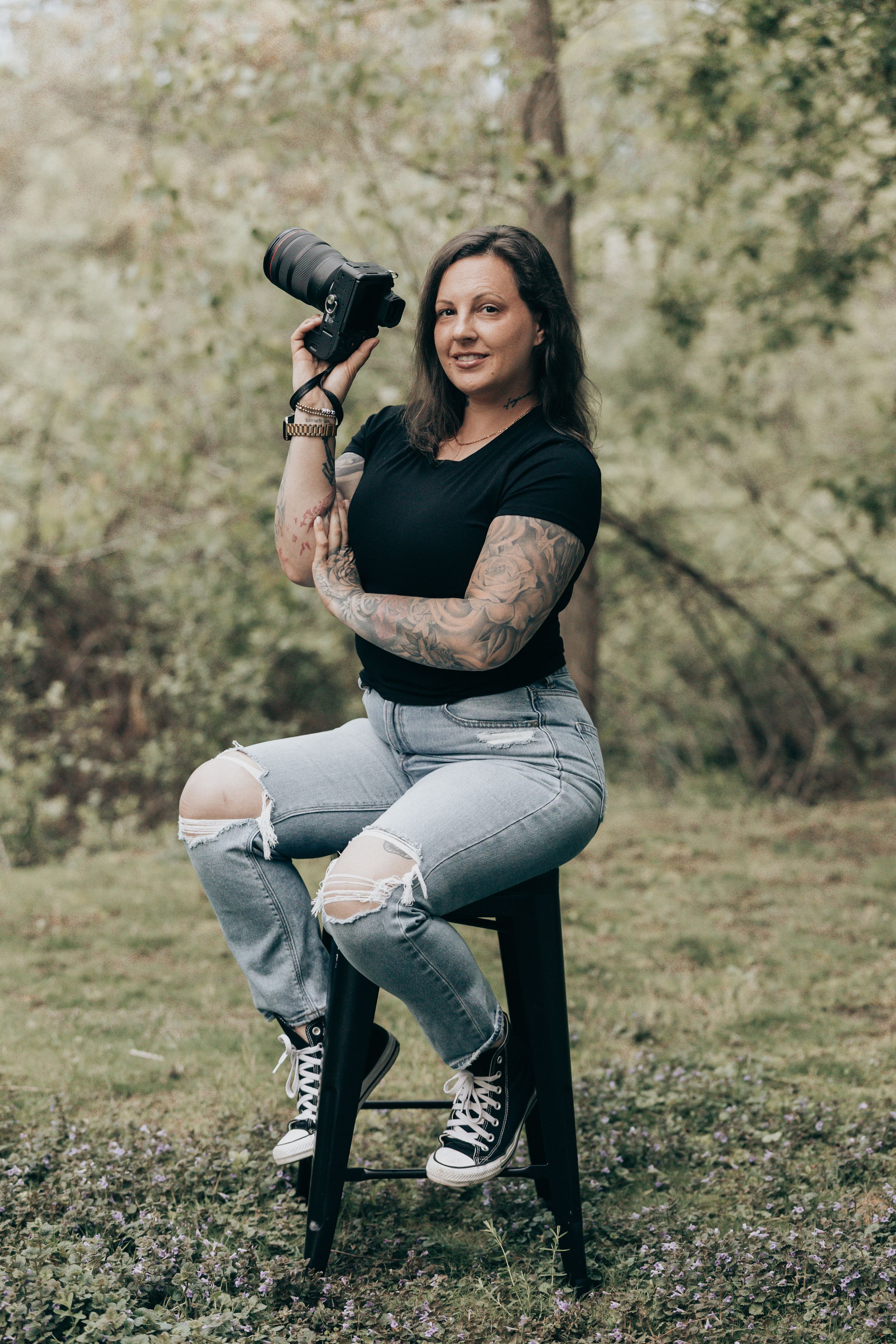 Photographer in ripped jeans and black shirt sits on stool outdoors holding professional camera in natural setting.