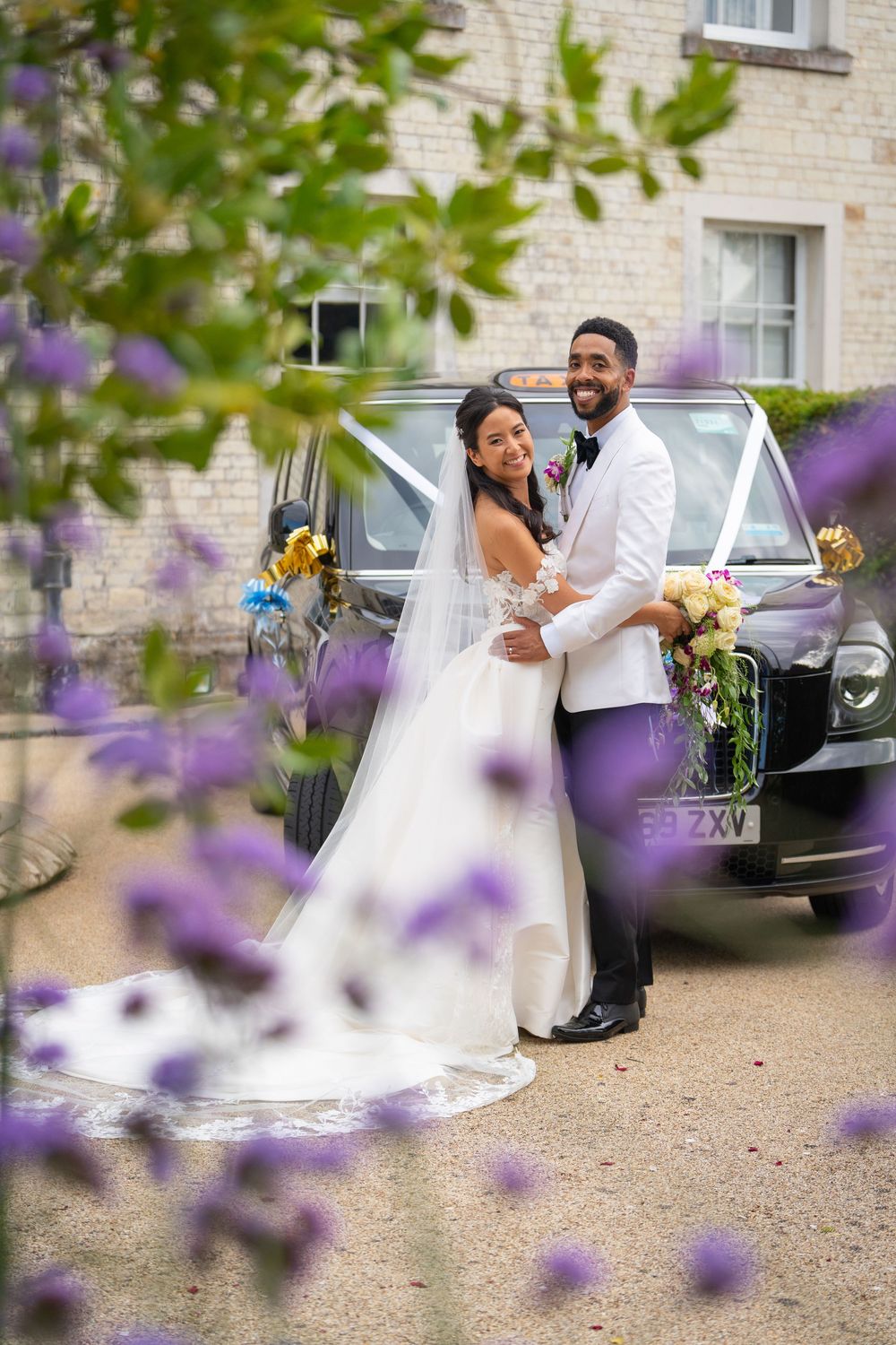 A wedding couple poses against a car through purple flowers on their special day at a stone building.