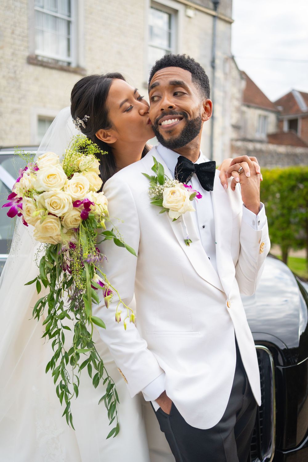 A joyful newlywed couple in matching white attire share a romantic moment with a beautiful bouquet of roses.