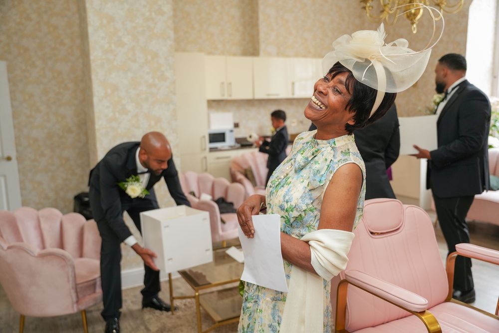 A person in a mint green dress and fascinator hat laughs joyfully while others unpack boxes in an elegant room.