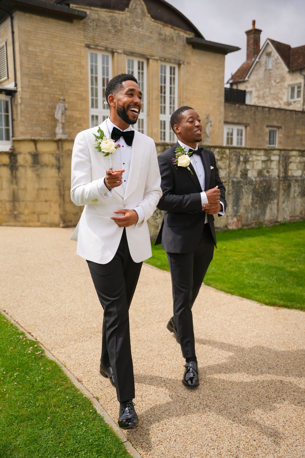 Two wedding guests in formal attire laugh while walking together on path outside historic stone building.
