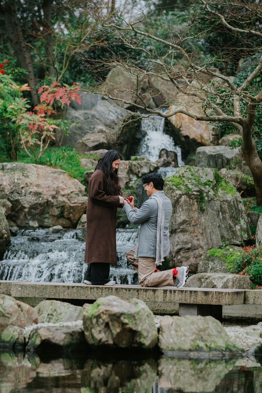 Two people share a romantic moment on a wooden bridge in front of a cascading waterfall surrounded by lush greenery.