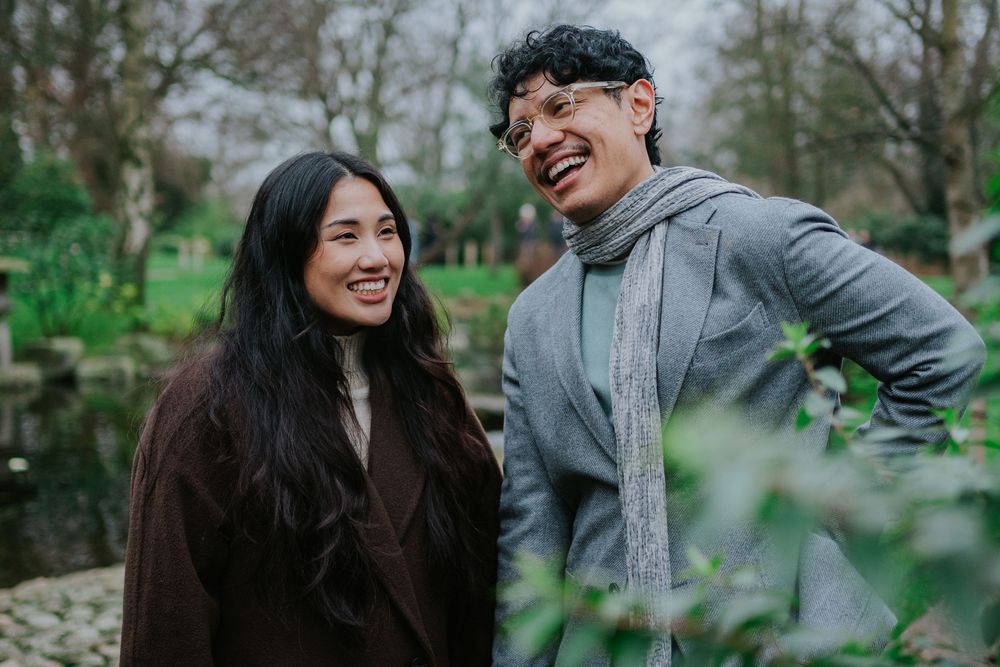 A young couple laughs together outdoors on a cloudy day with greenery in the background.