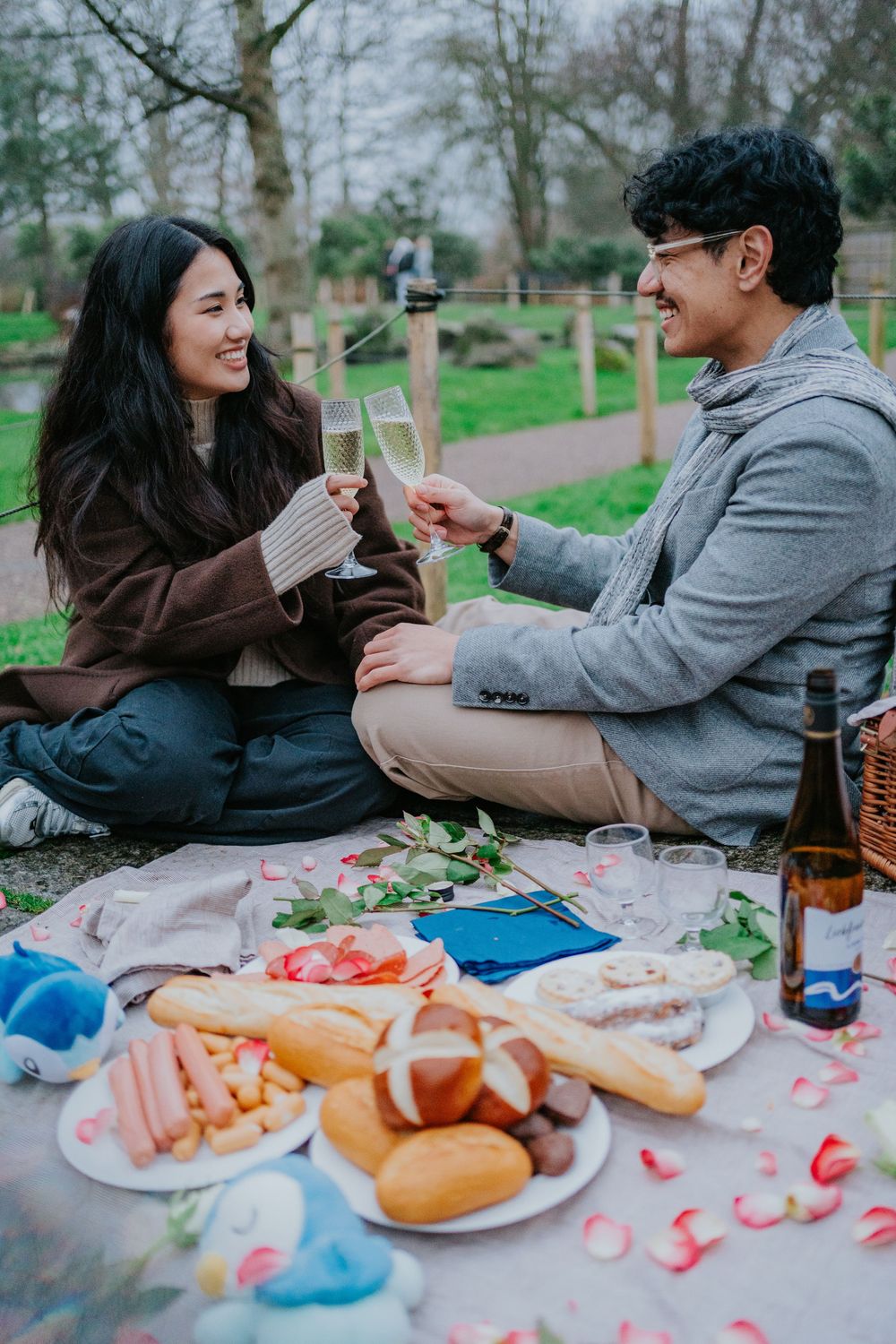 Couple enjoying romantic outdoor picnic with sandwiches and drinks while clinking glasses together in a park setting.
