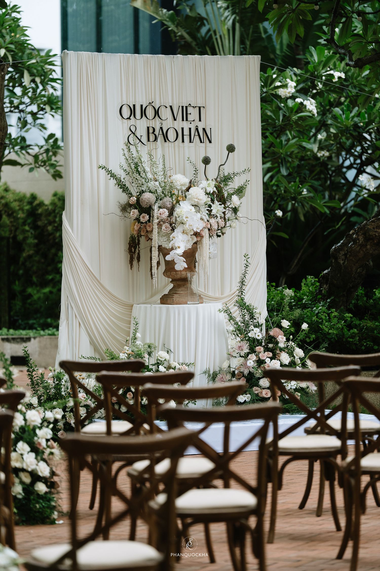 Romantic outdoor wedding ceremony setup with cream drape backdrop, floral arrangements, and wooden cross-back chairs.