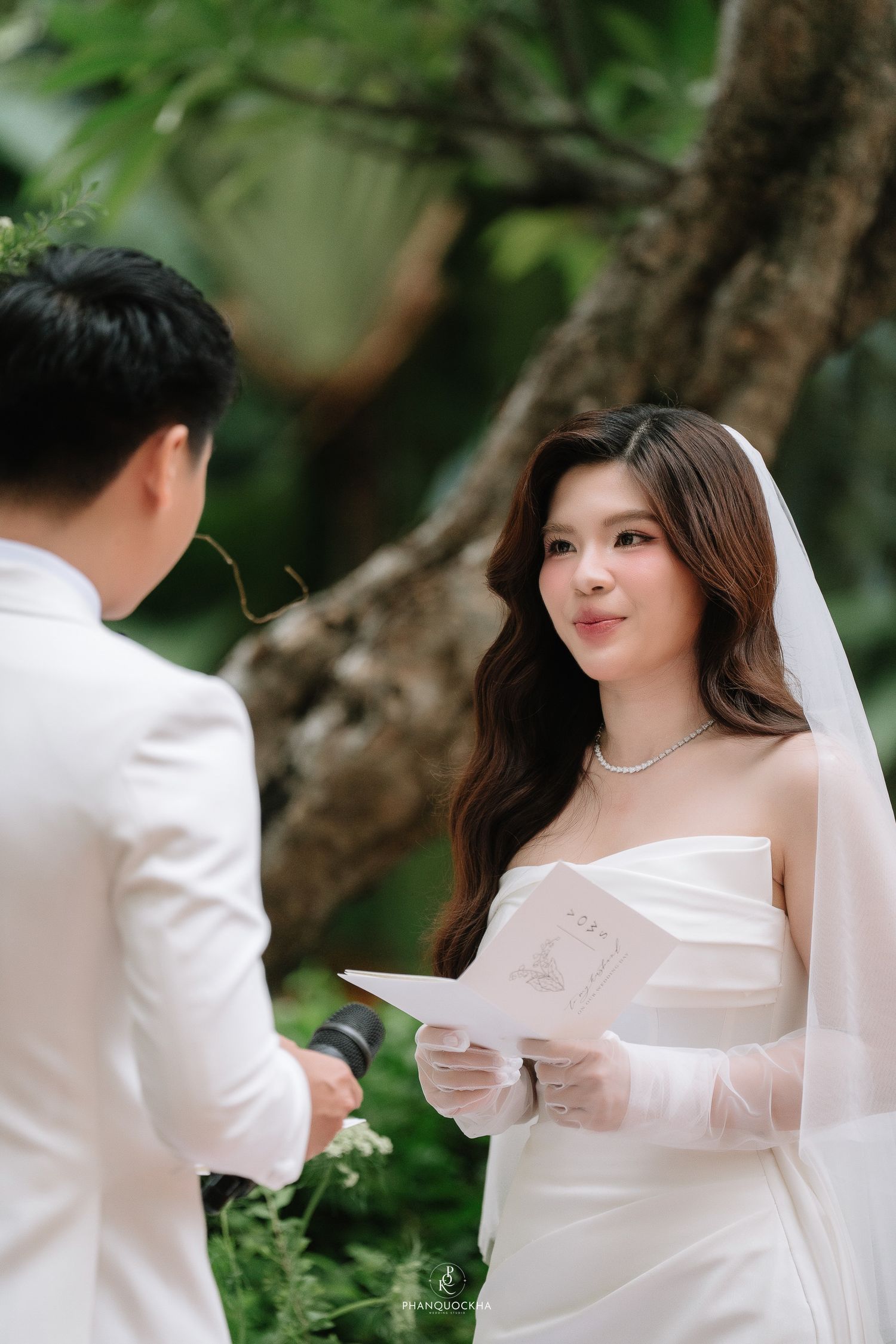 Beautiful bride in strapless white gown and veil reads wedding vows while gazing at groom outdoors.