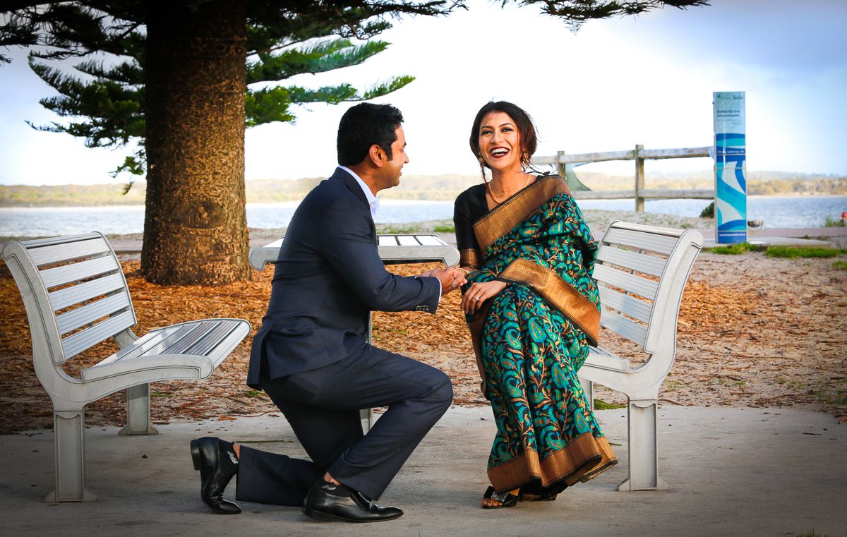 Romantic proposal scene at sunset on beach with couple sitting on white wooden bench under pine trees.