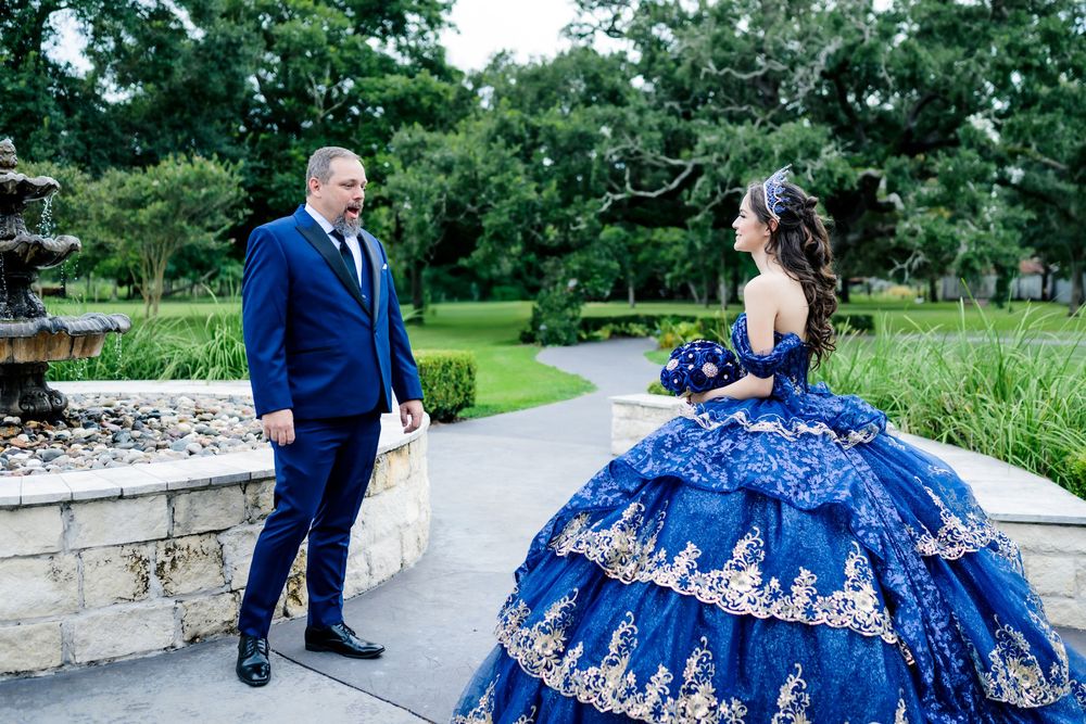 A couple in formal attire, with a magnificent deep blue ballgown with silver embellishments, pose in a garden setting.