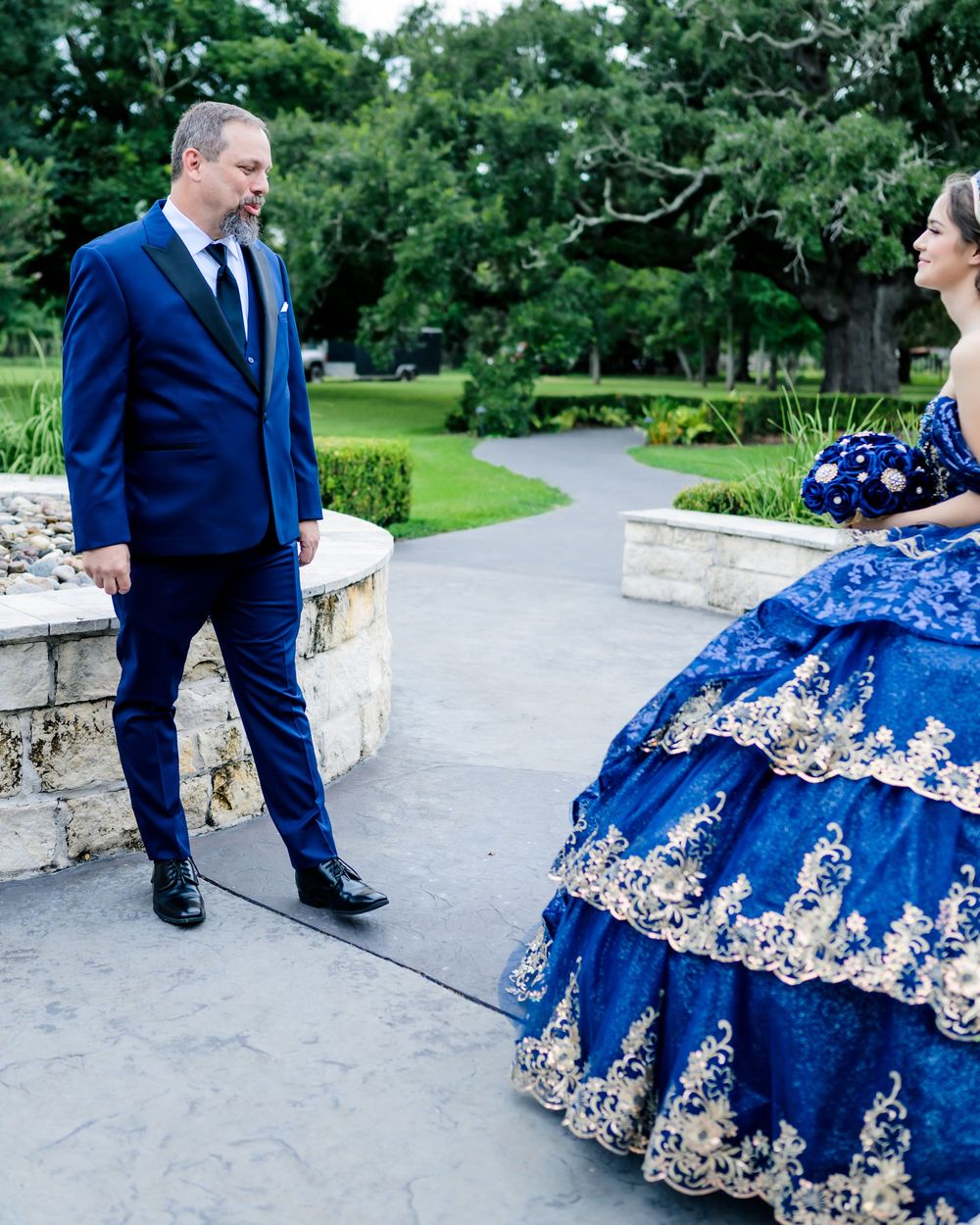 A formal couple in coordinating royal blue attire pose on a garden path with elegant stone walls.