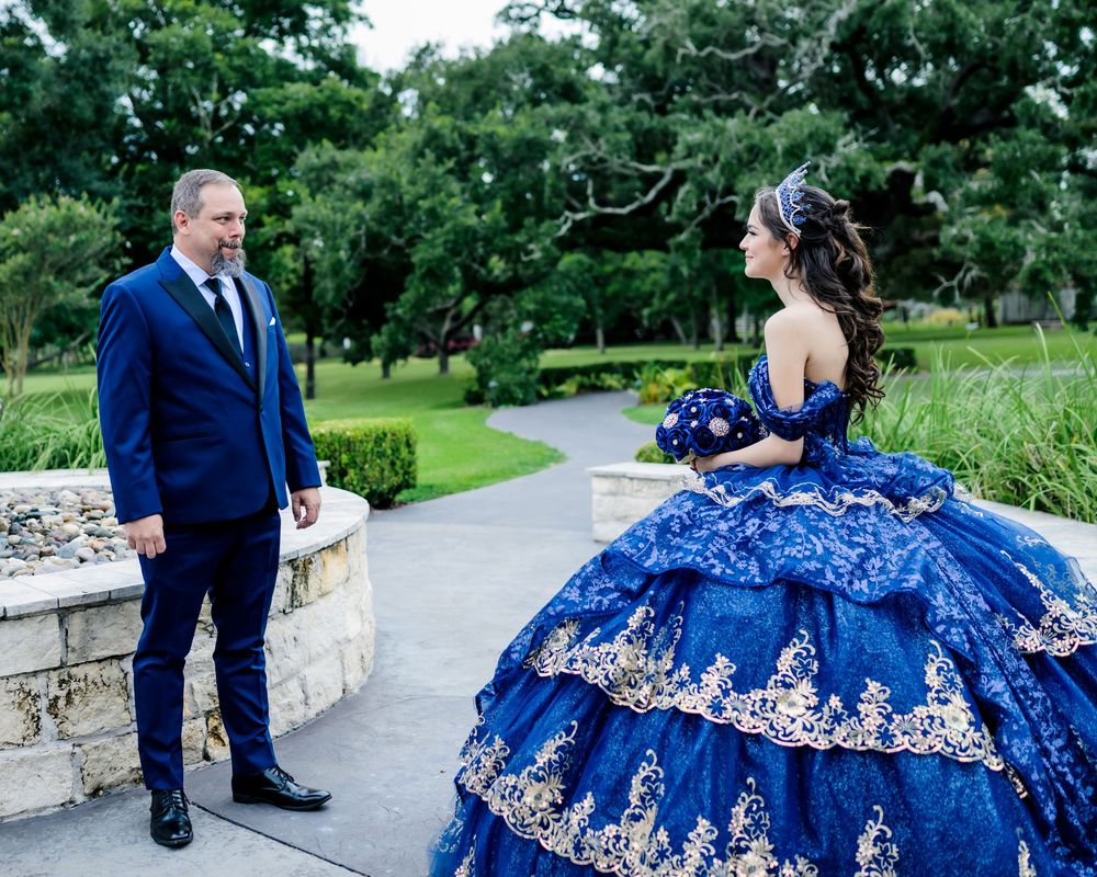 A couple in formal attire, with a stunning royal blue quinceañera dress with silver embroidery standing on a garden path.