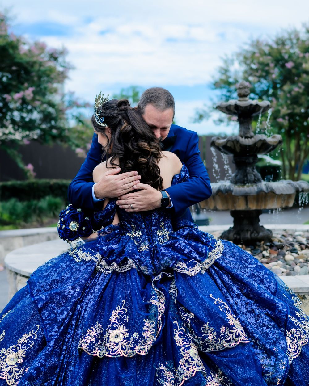 A black dog being embraced in front of an ornate fountain while resting on a beautiful royal blue and silver embroidered gown.