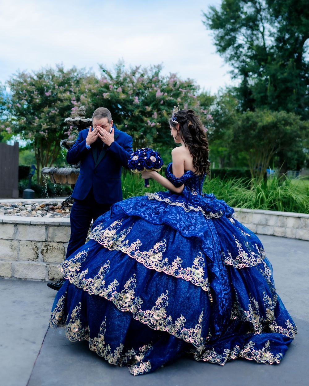 Two people dressed in elegant royal blue attire pose outdoors against a garden backdrop during a formal celebration.