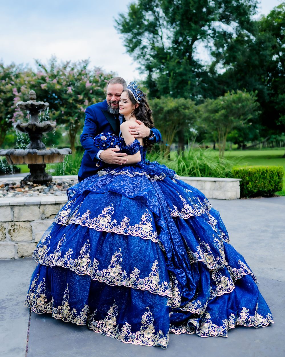 A couple embraces while wearing matching blue formal attire in a garden setting with a decorative fountain.