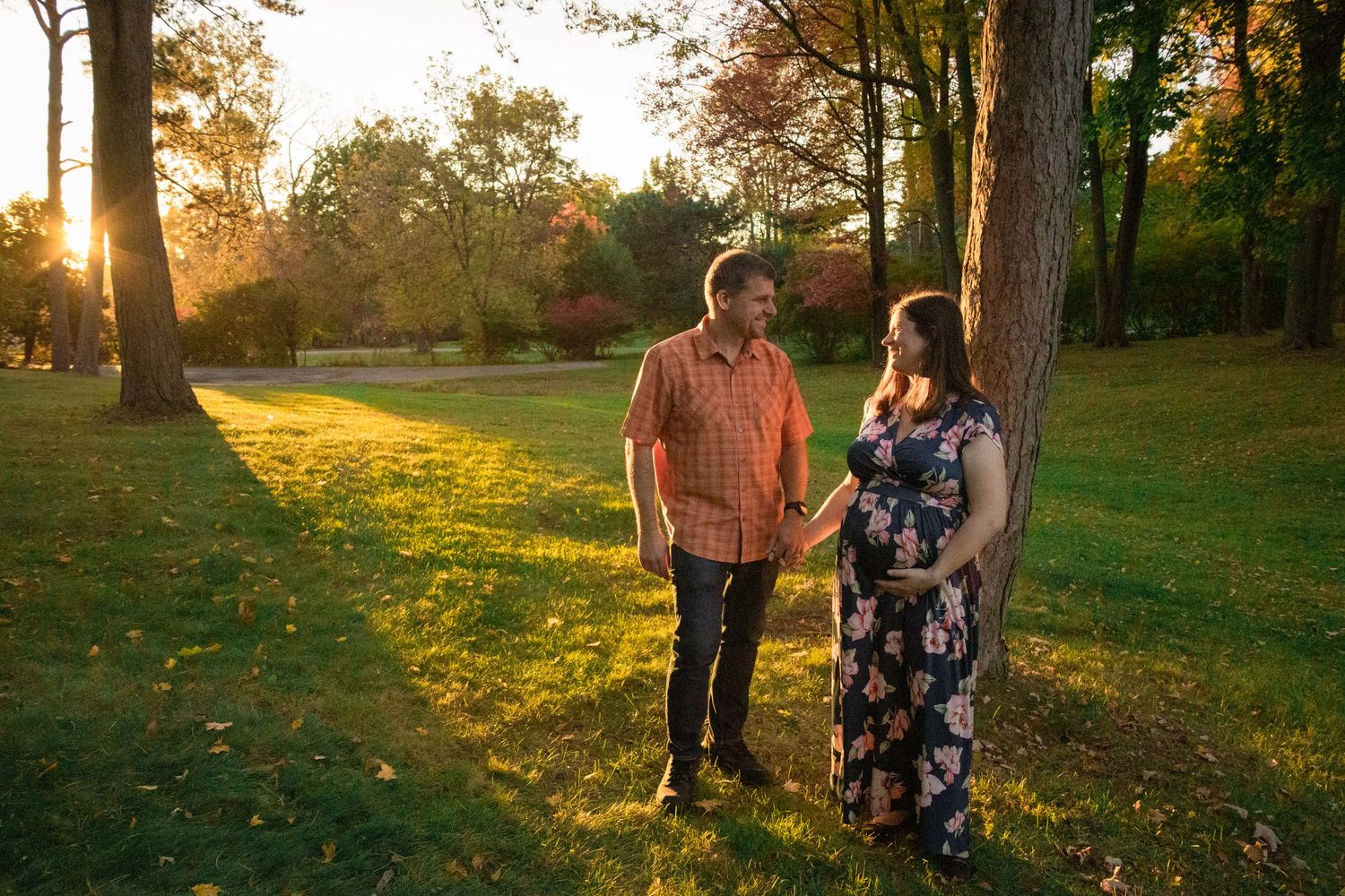 Expecting couple shares tender moment in golden sunset light while wearing floral dress and plaid shirt.