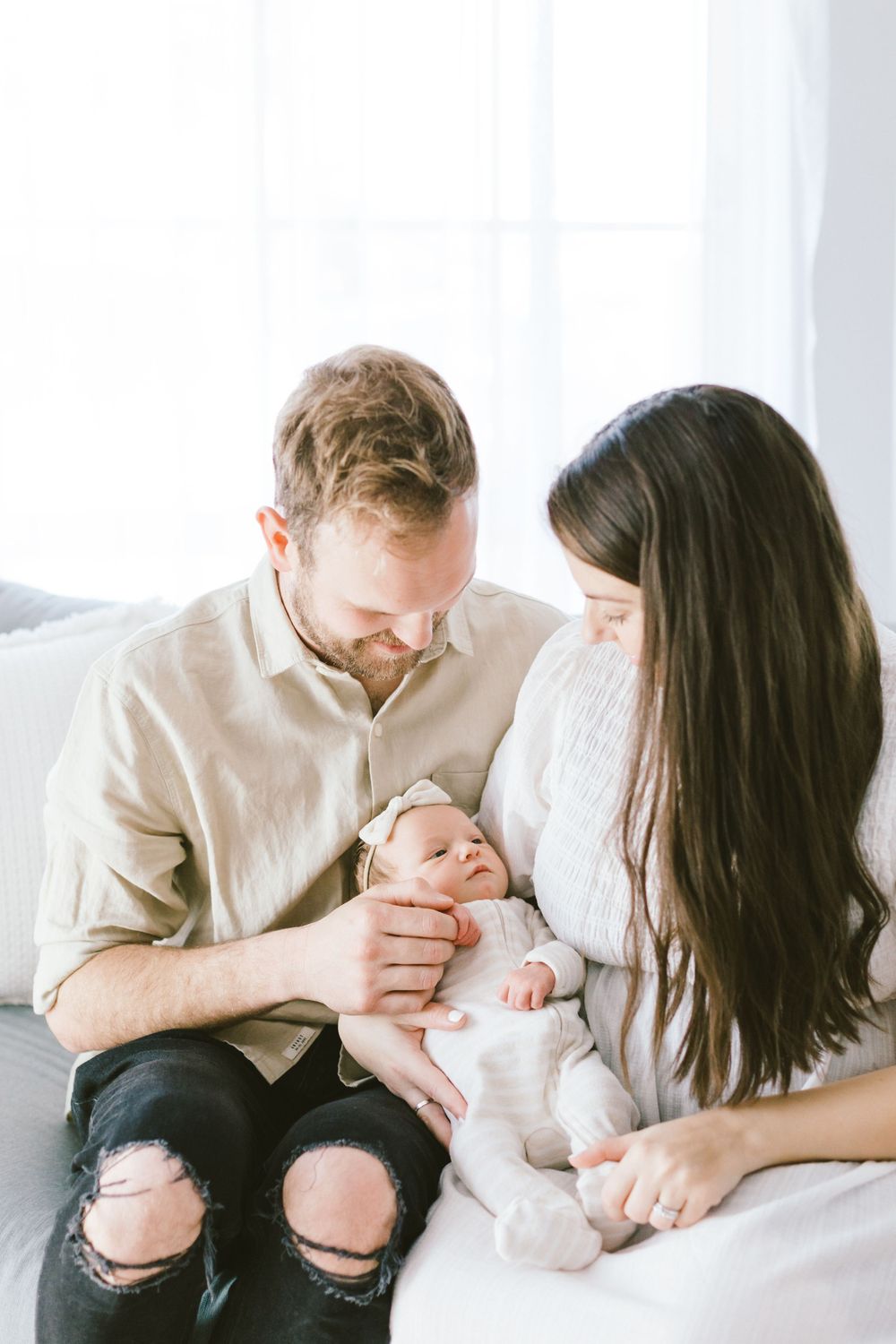 Family cuddling with newborn baby dressed in white against bright window.