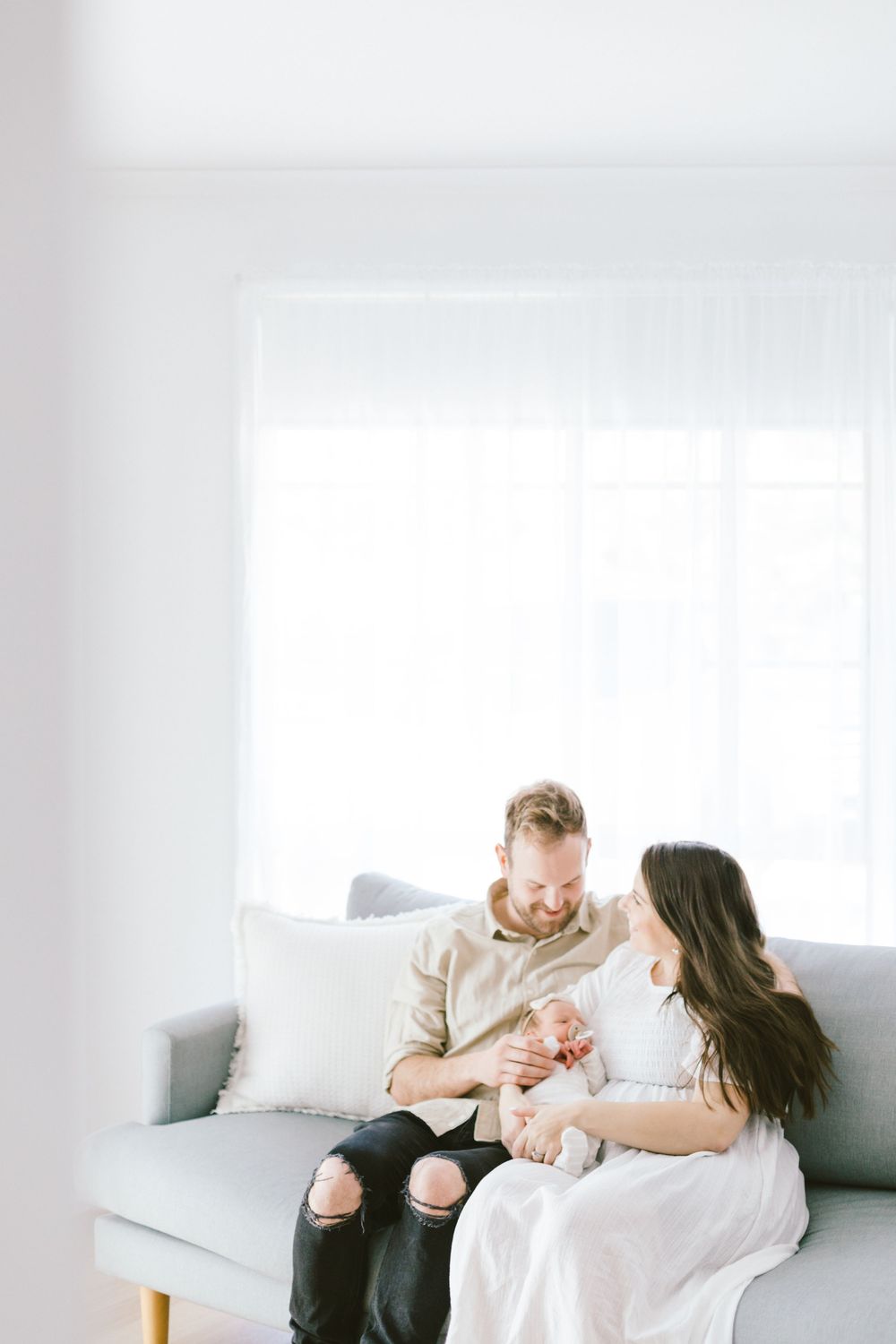 A couple relaxes on a white couch while cuddling their newborn baby in a bright minimalist room setting.