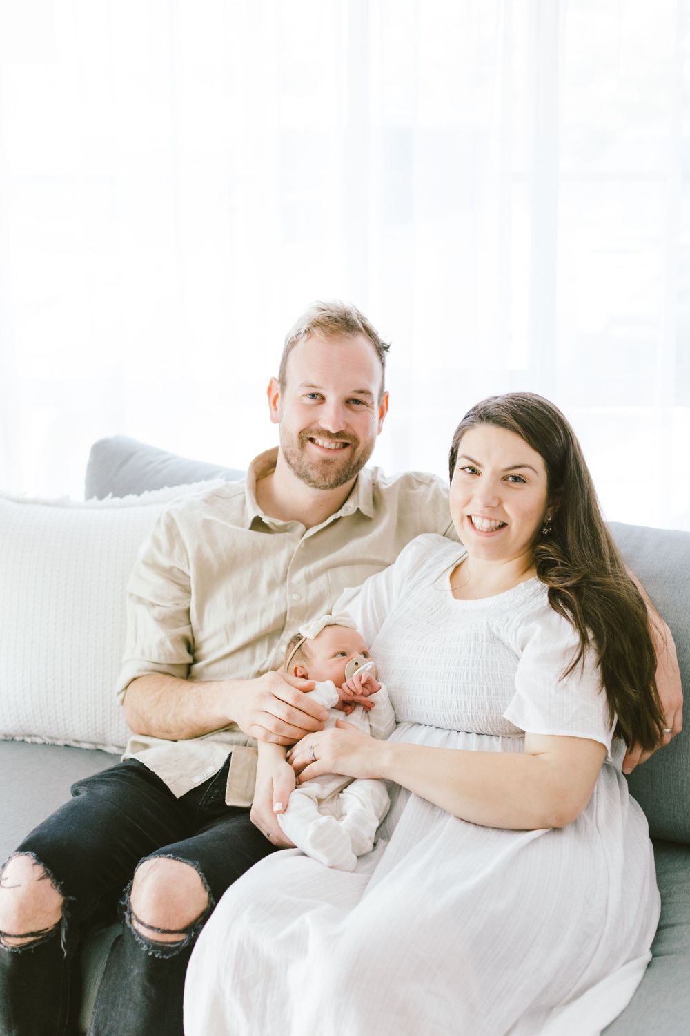 Family sits together on white sofa with their newborn baby during an intimate lifestyle photoshoot.