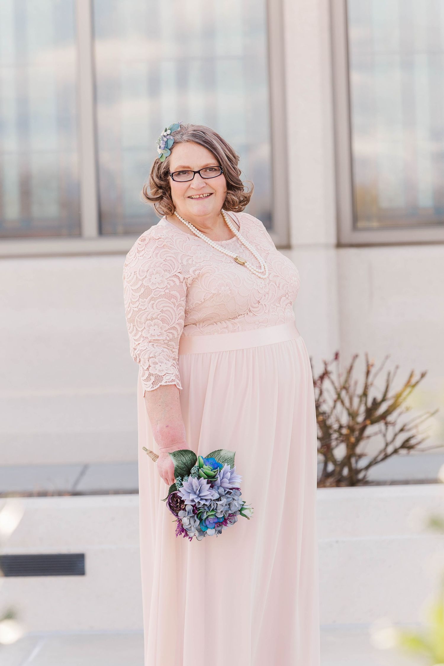 Woman in pink lace dress holding blue flowers against light building exterior.
