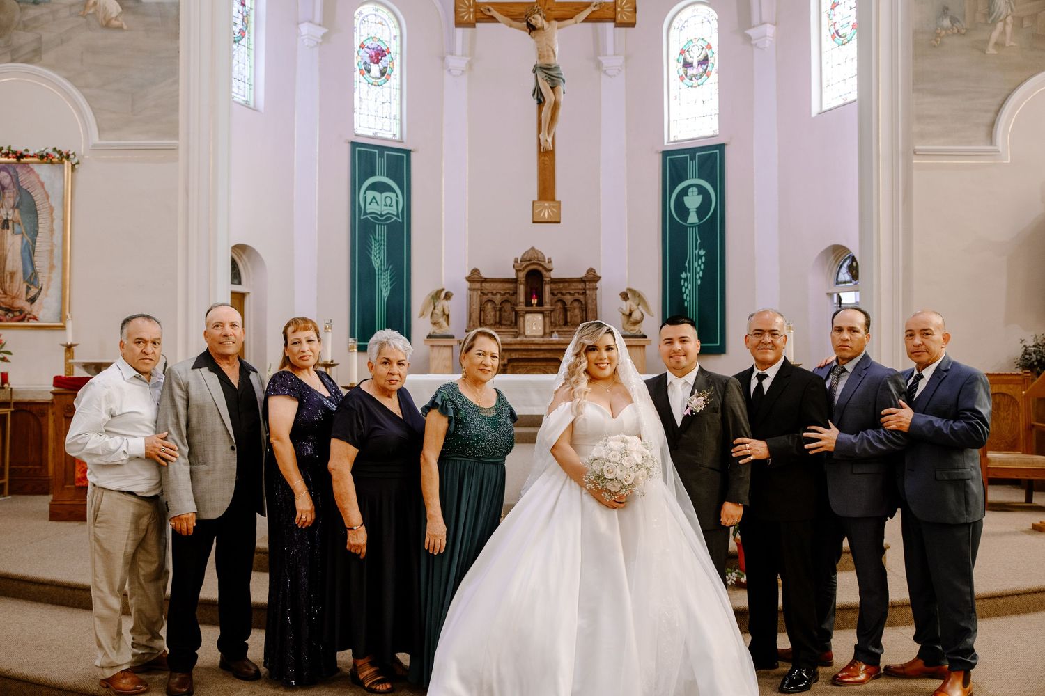 A wedding party poses together inside a beautiful white Catholic church with stained glass windows and a crucifix altar.