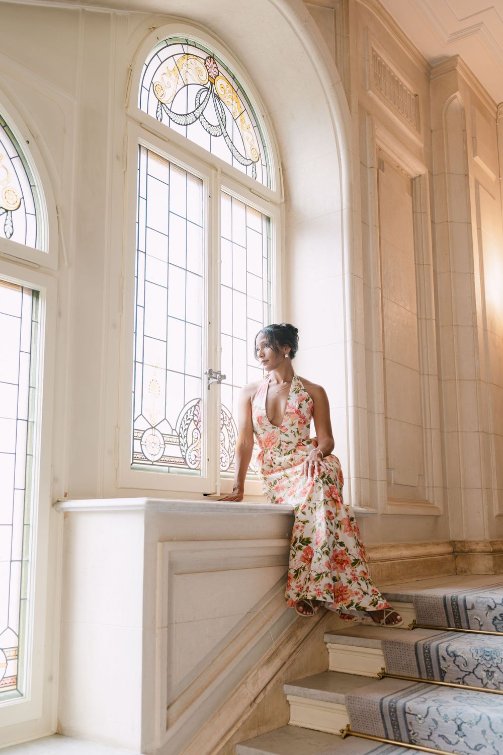A floral dress flowing down marble stairs in front of an elegant arched window with intricate stained glass.