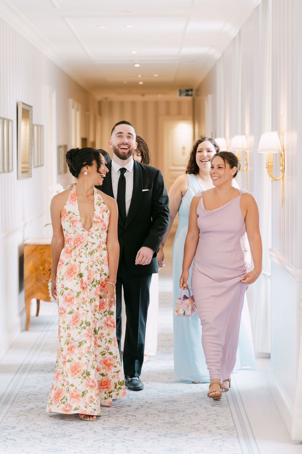 Wedding party walks and laughs together in an elegant hotel hallway with floral dresses and formal attire.