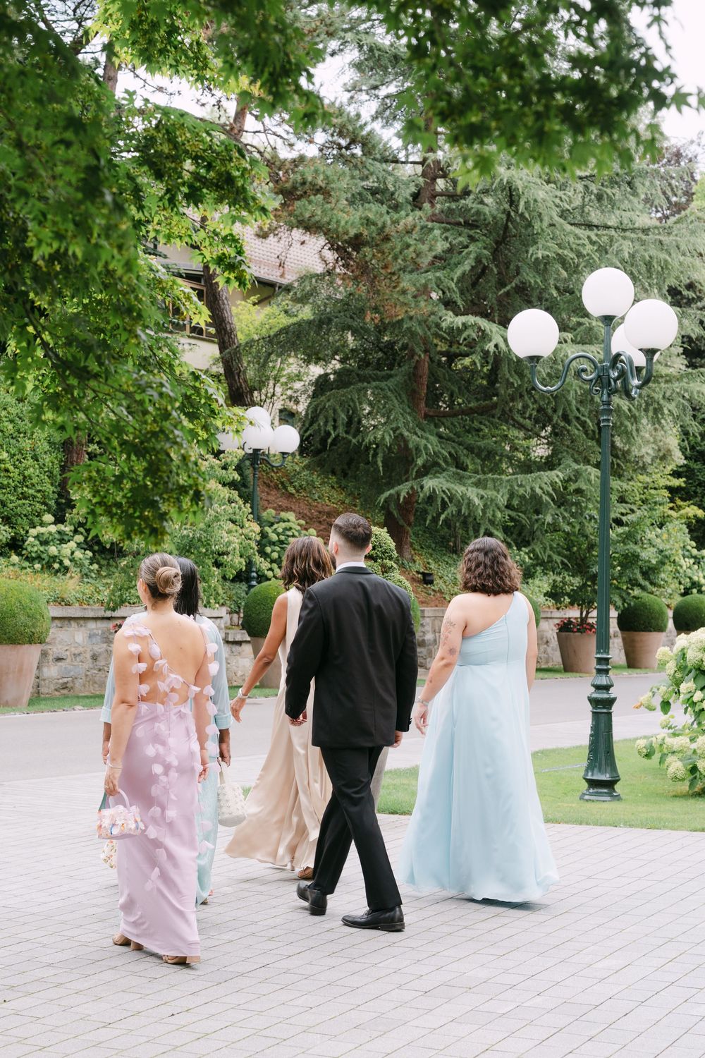 Wedding party members in pastel dresses and formal attire walk along a garden path lined with ornate lampposts.