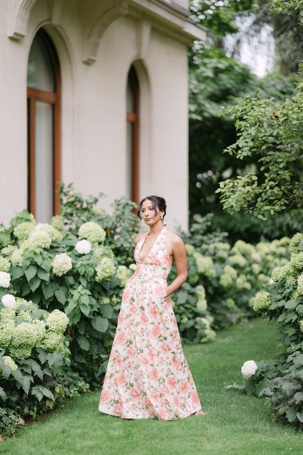 A floral print summer dress stands out beautifully against a garden backdrop with white hydrangea bushes and classic architecture.